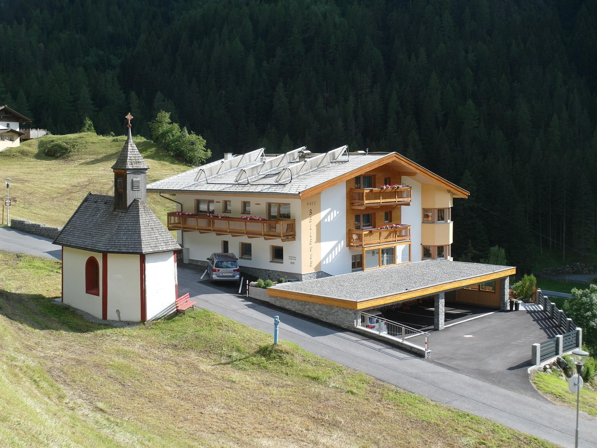 A modern building with balconies stands next to a small chapel in a mountainous area. In the background, green forests can be seen.