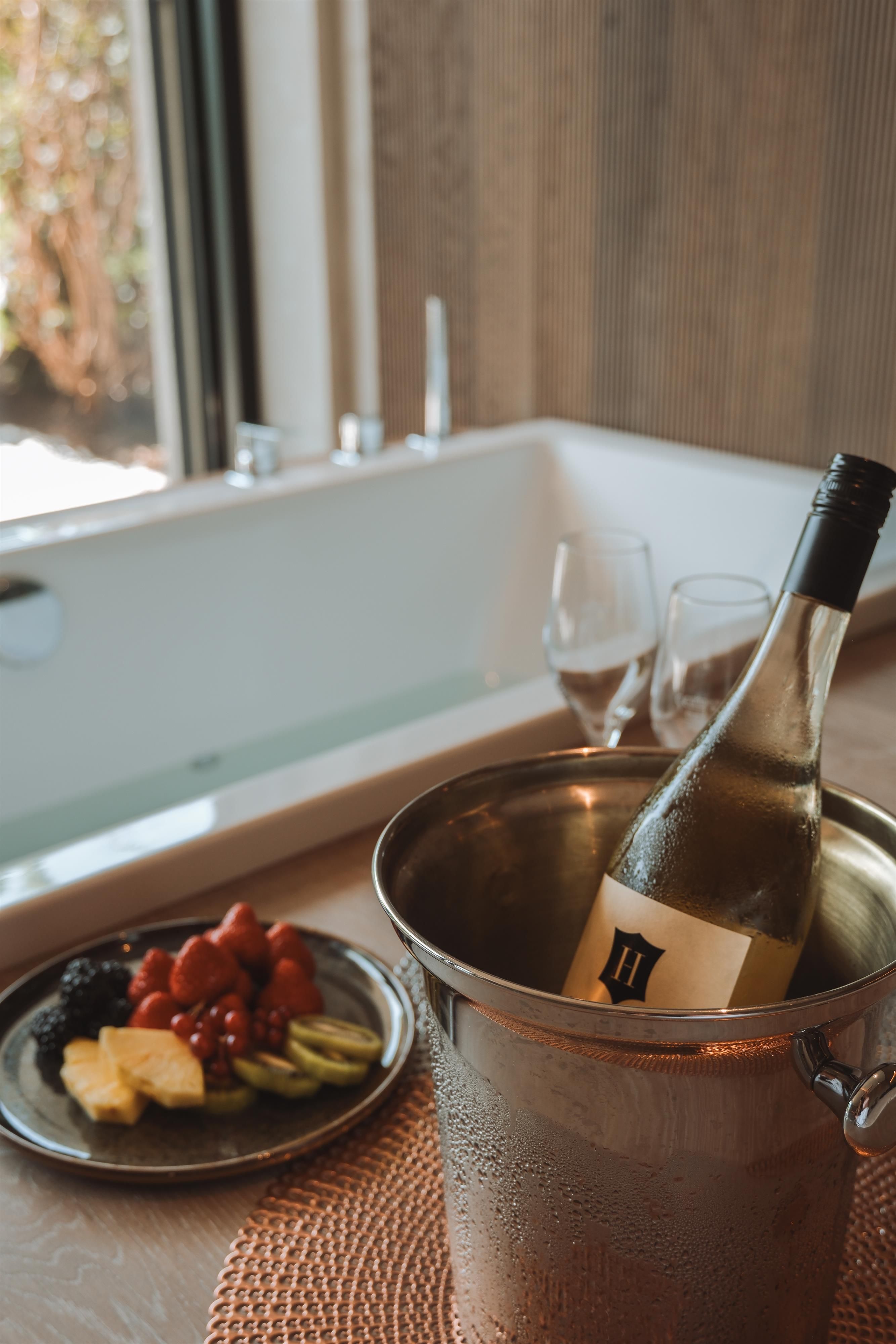 An elegant table with an ice bucket containing a bottle of white wine. Next to the bucket are fresh fruits and two wine glasses, while a bathtub is visible in the background.