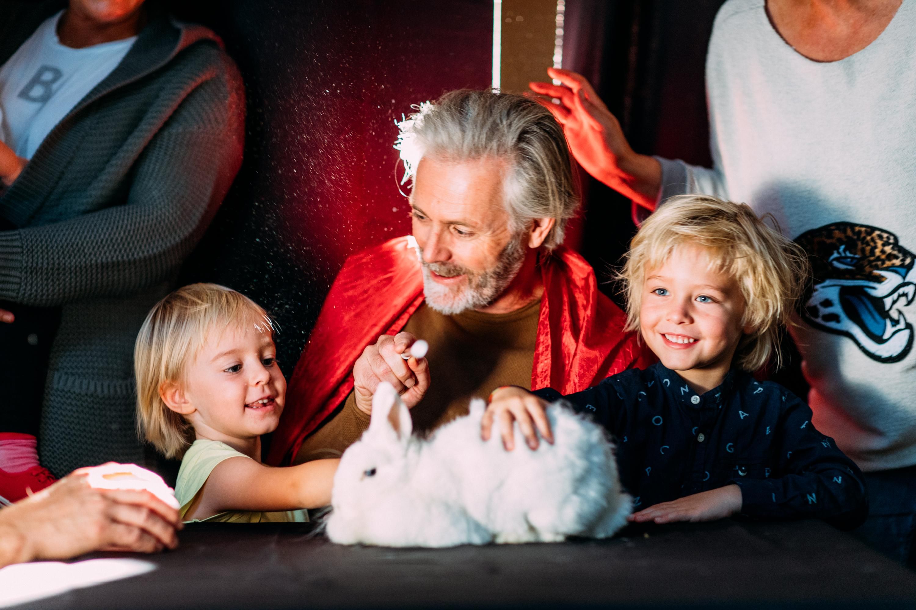 An older man with gray hair is sitting at the table holding a white rabbit. Children are standing around him, enjoying the animal.