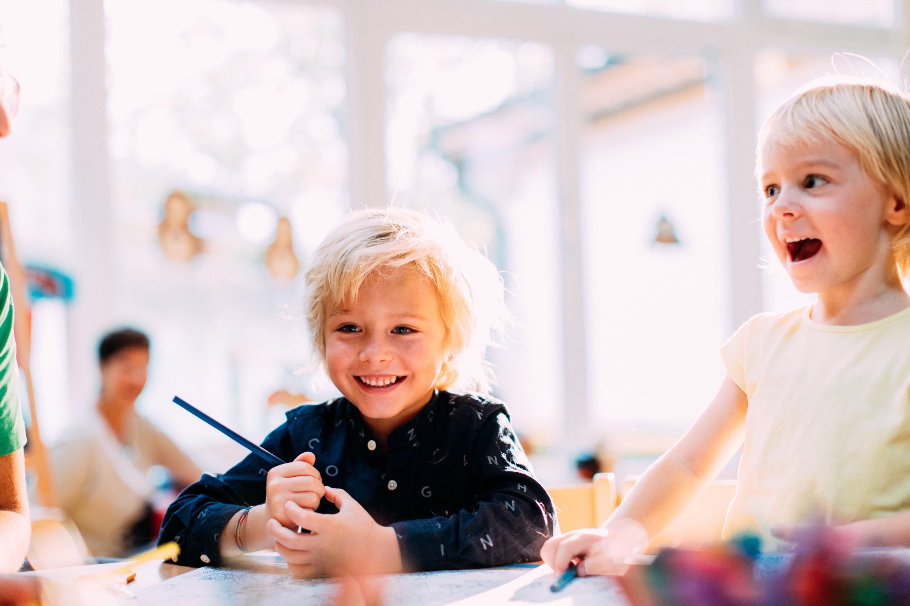 Two cheerful children are sitting at a table and laughing. They are holding colored pencils in their hands and seem to be creative.