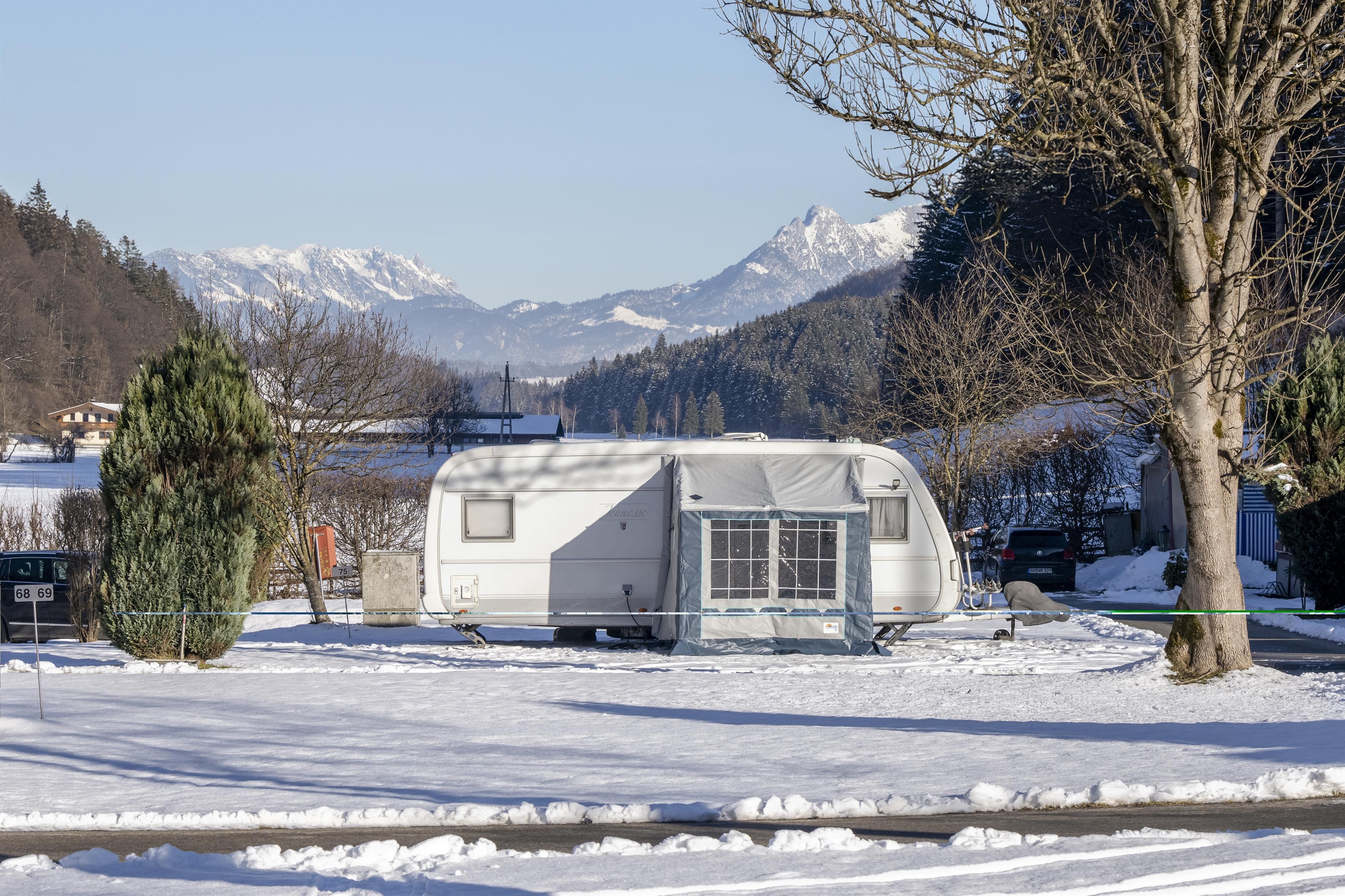 A caravan stands in a snow-covered landscape. In the background, there are snow-covered mountains and a blue sky.