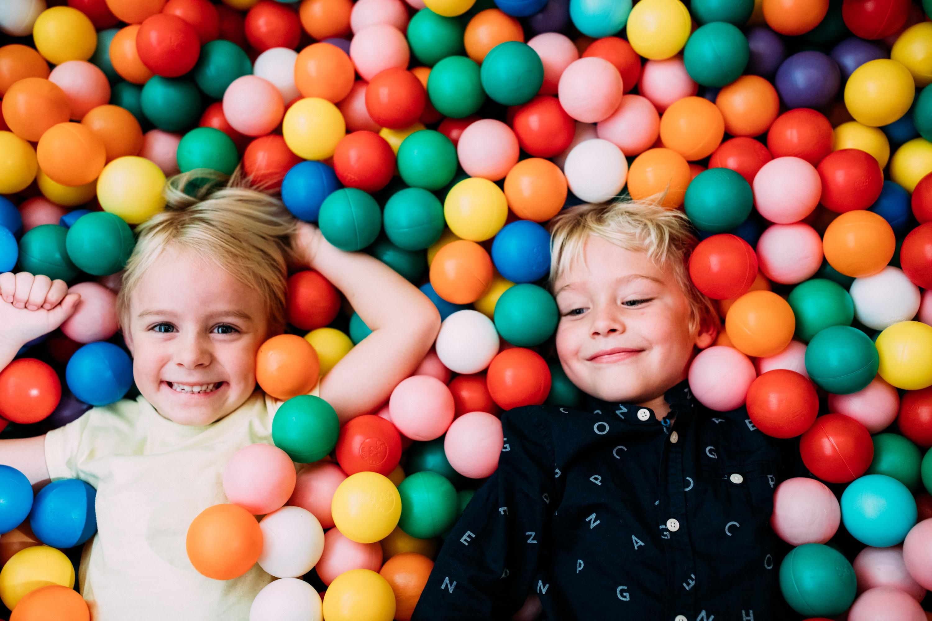 Two children are lying smiling in a colorful ball pit. Around them are many colorful plastic balls in various colors.