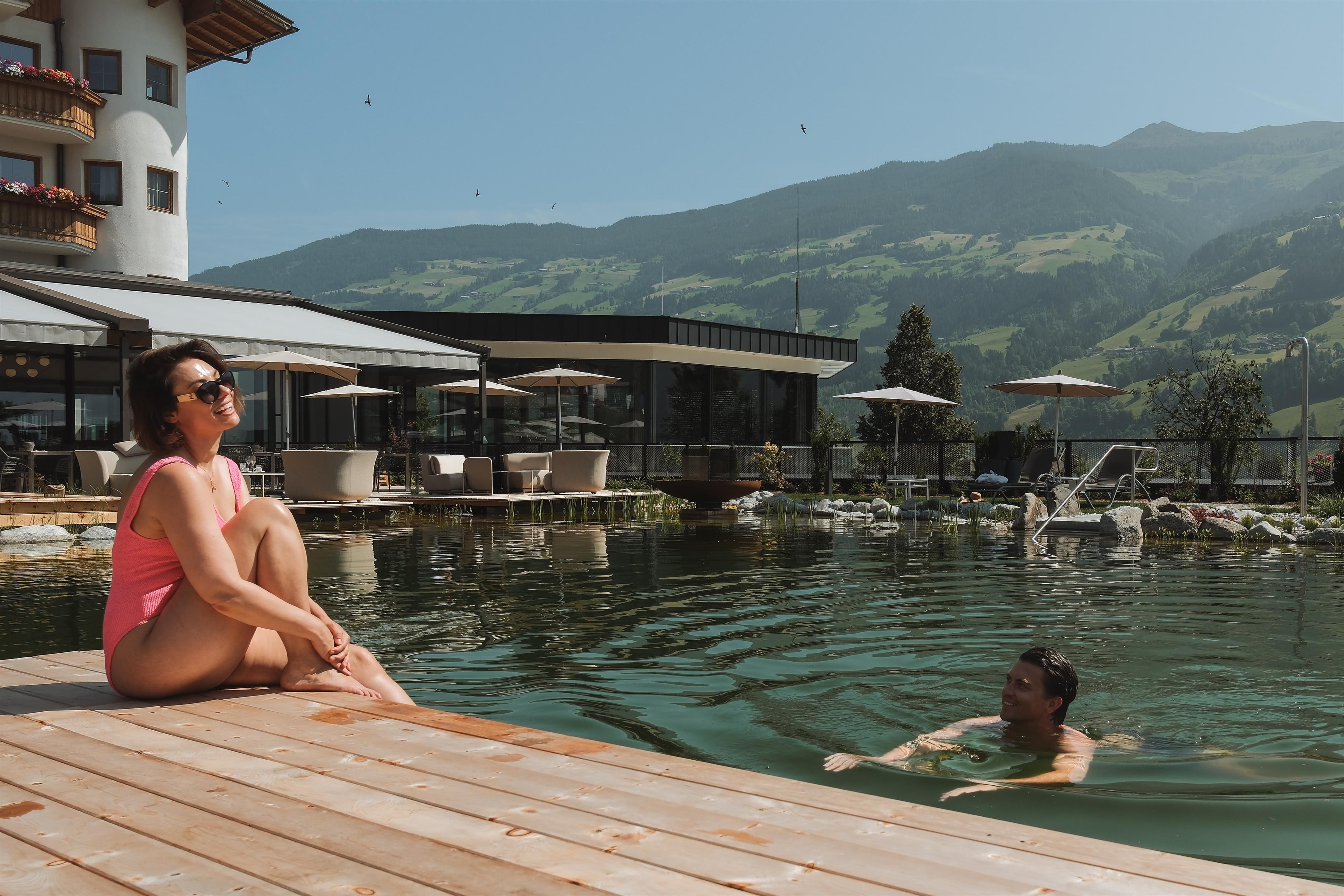 A pool in the mountains with a woman sitting by the edge, enjoying the sun. A man is swimming in the water, and in the background, there is a beautiful landscape and a hotel visible.
