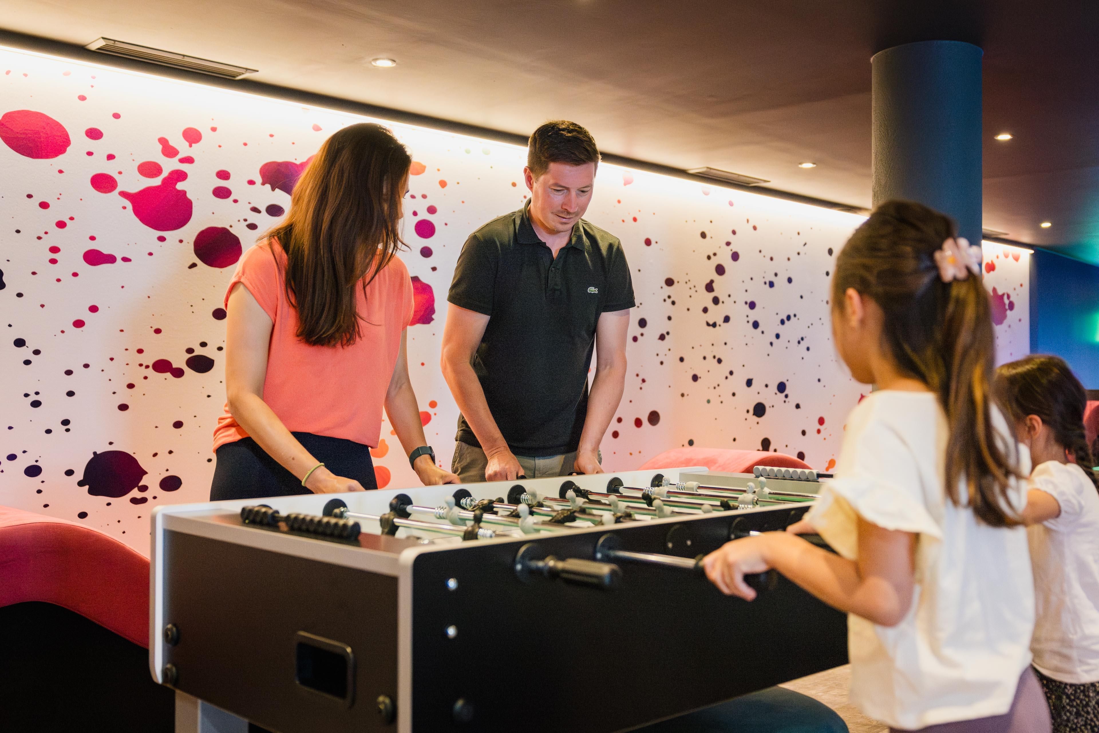 Two adults and two children are playing a foosball duel in the new play area for the whole family. In the background, there is a colorful wall with splashing colors.
