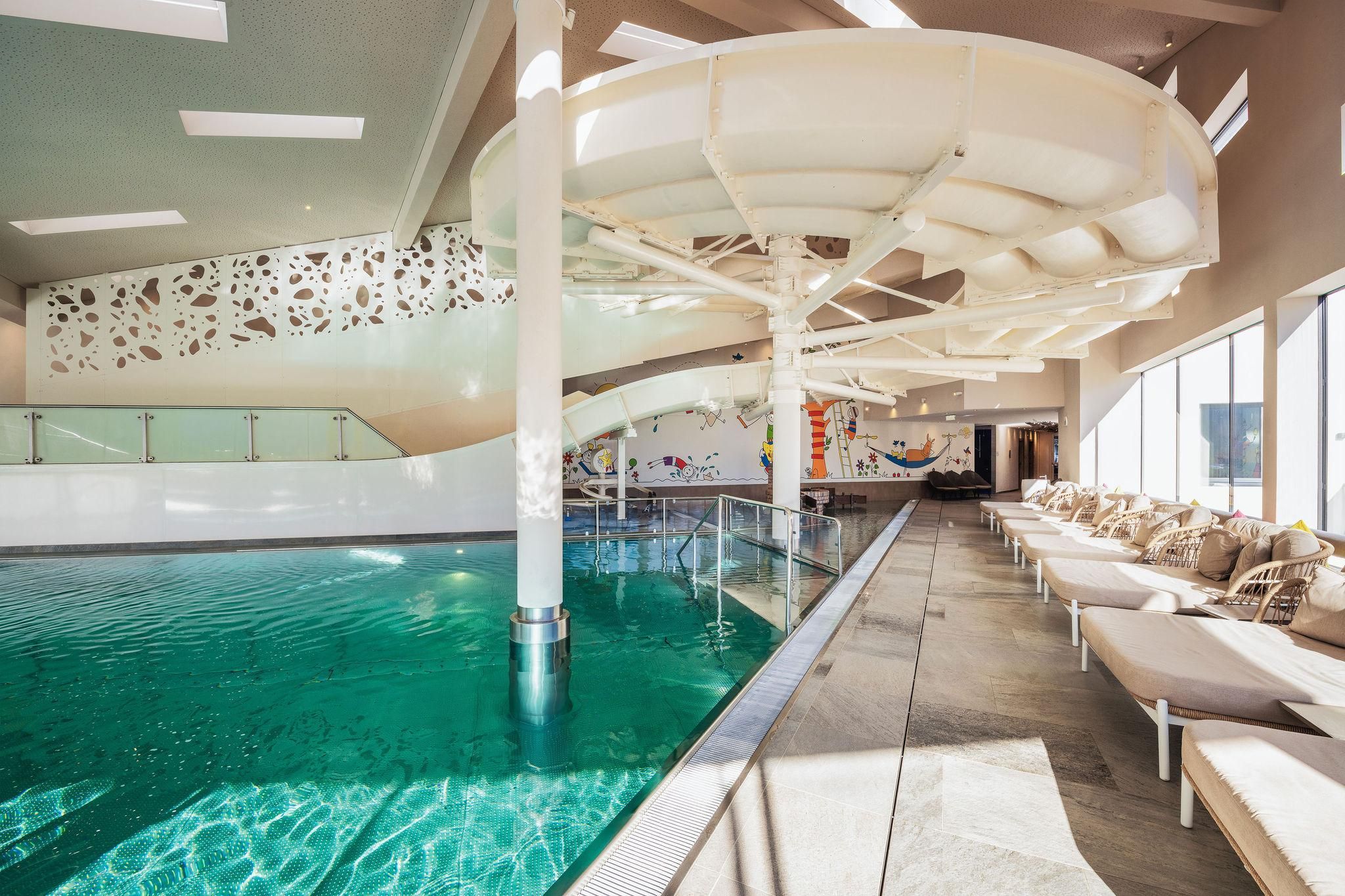 A modern swimming pool with a slide and comfortable loungers. The well-lit room features large windows and an inviting atmosphere.