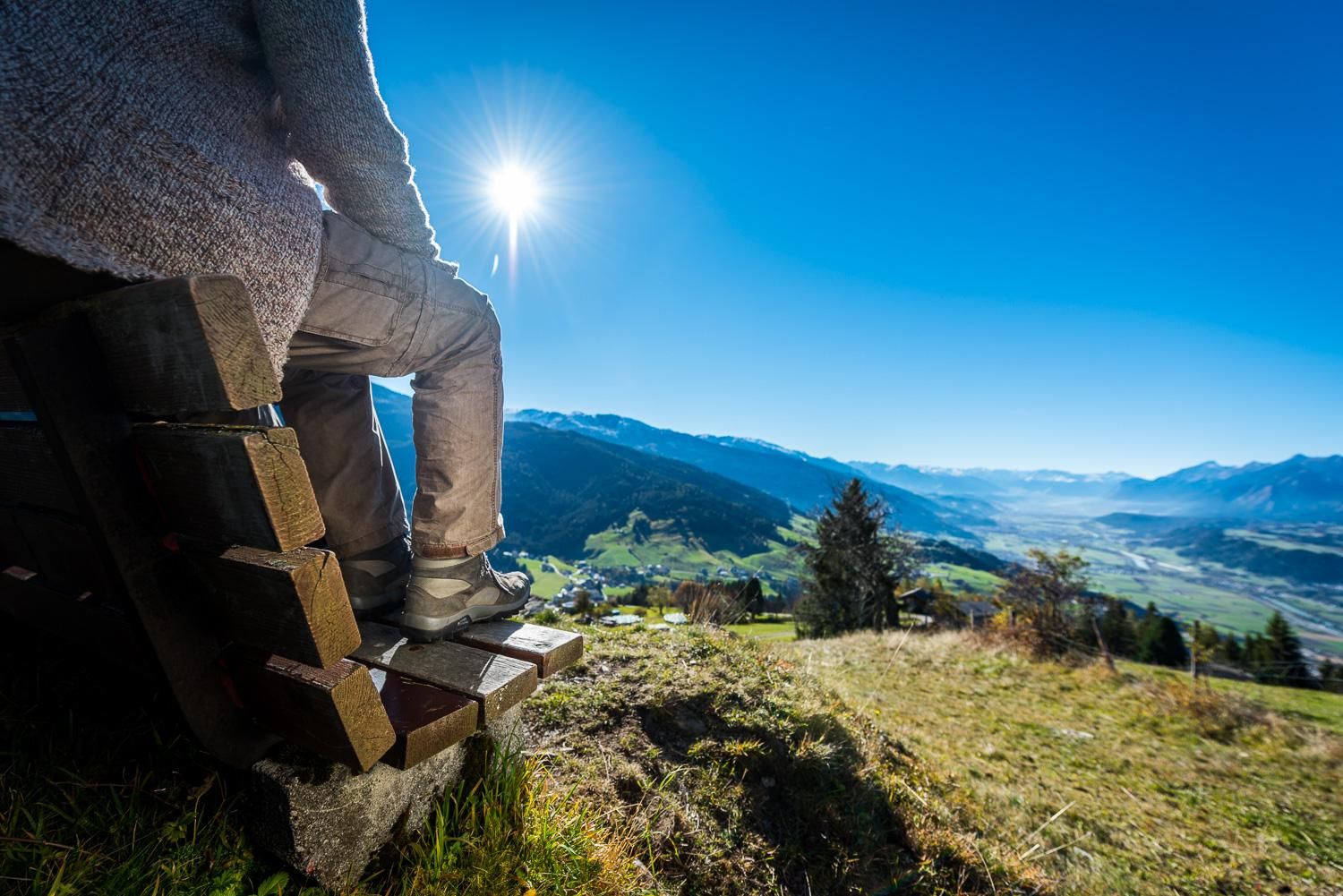A person is sitting on a bench overlooking the valley. The sun is shining in the clear blue sky and the landscape is green and hilly.
