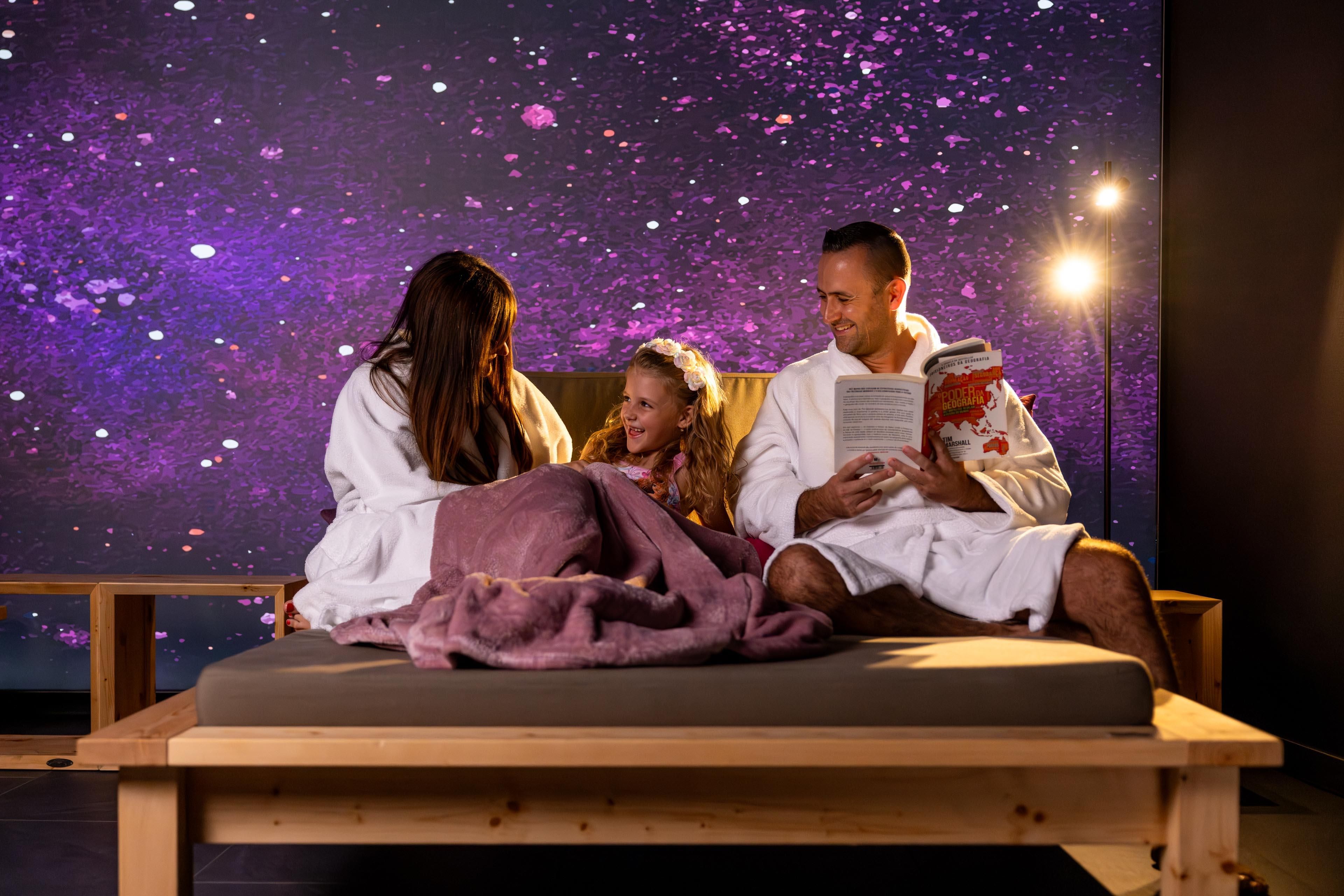 A family is comfortably seated on a lounge in the relaxation room, enjoying a relaxed time together. A galactic background is visible, creating a soothing atmosphere.