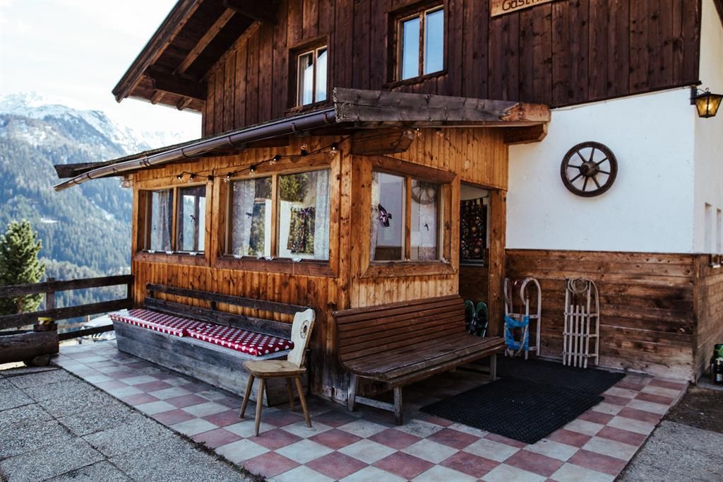 A cozy wooden mountain cabin with a covered area and windows. In the background, the mountains are visible, and the floor is designed with colorful tiles.