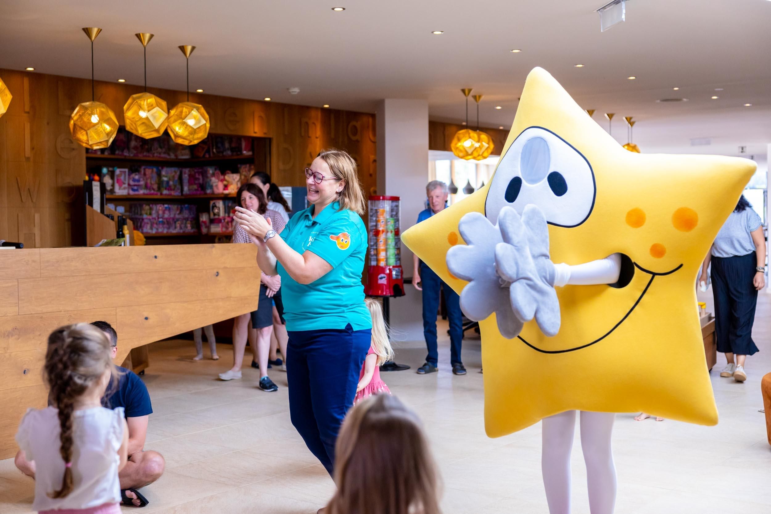 The children are dancing with the caretaker and the mascot Laurentius, a large yellow star, in the hotel lobby and are having a lot of fun.