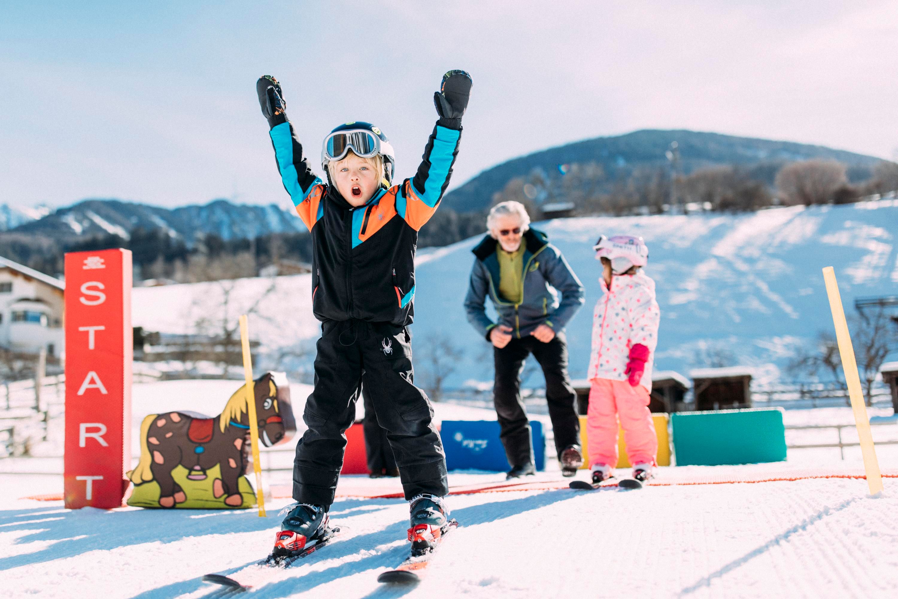 A happy child is skiing and cheering at the starting point. In the background, an adult and another child can be seen, along with a snowy landscape.