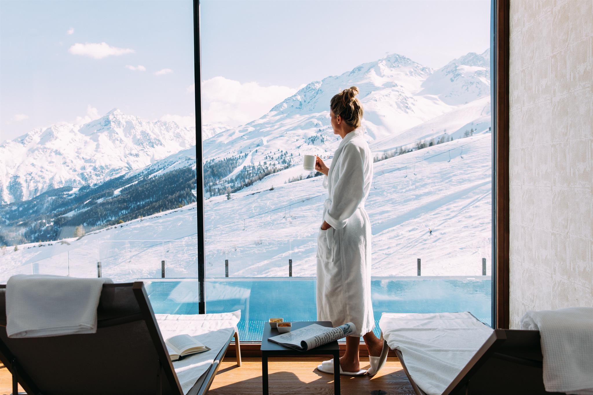 A woman in a bathrobe stands in front of a large window and looks at snow-covered mountains. The view conveys tranquility and relaxation.