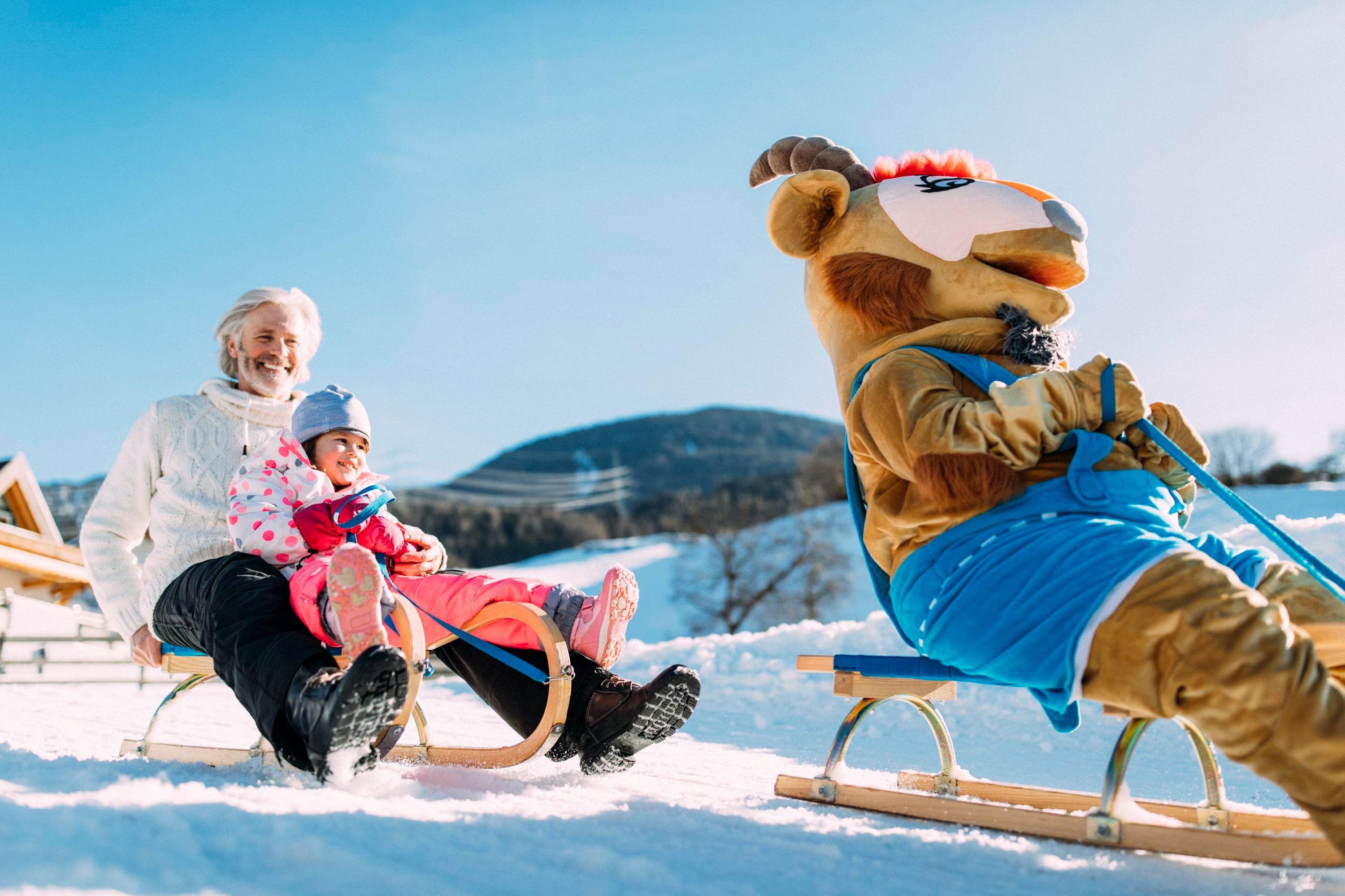 A grandfather with his grandchild sits on a sled in the snow. Next to them is a mascot joyfully riding on a sled.