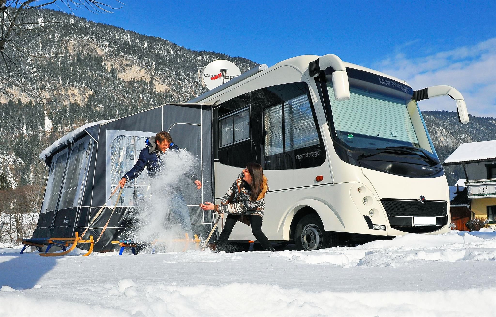A motorhome is standing in the snow, surrounded by mountains. Two people are playing in the snow and having fun.