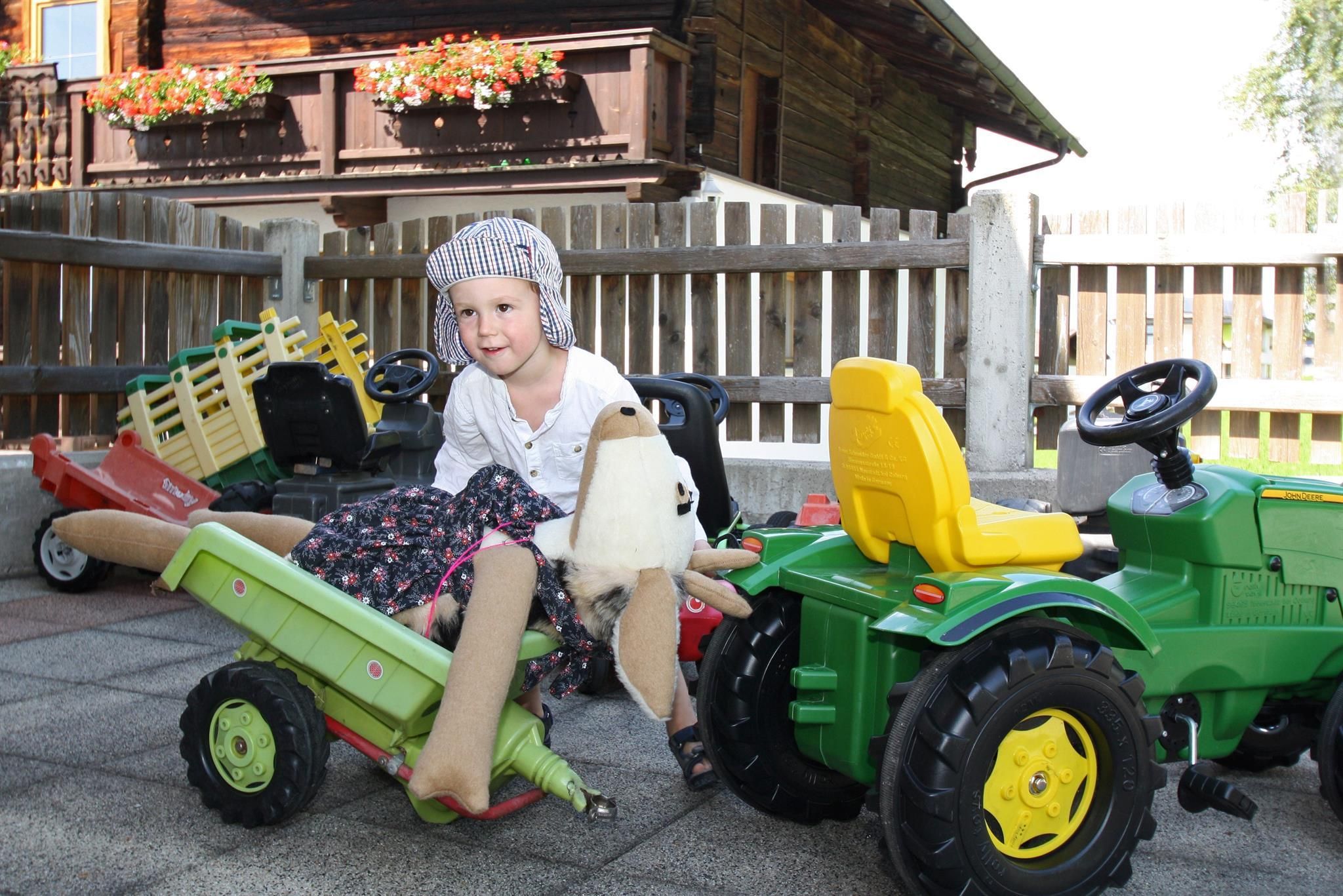 A small child is sitting on a toy tractor and smiling. In the background, there are more toys and a rustic wooden house visible.