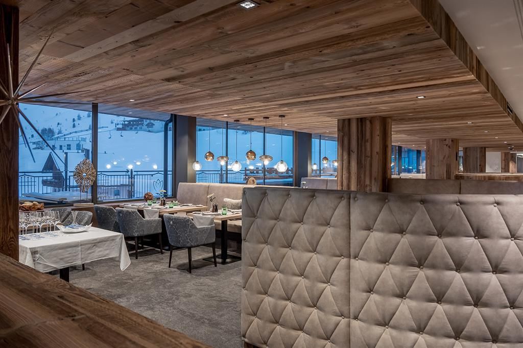A modern restaurant with wooden decor and cozy seating areas. Large windows offer a view of the snow and the mountains.