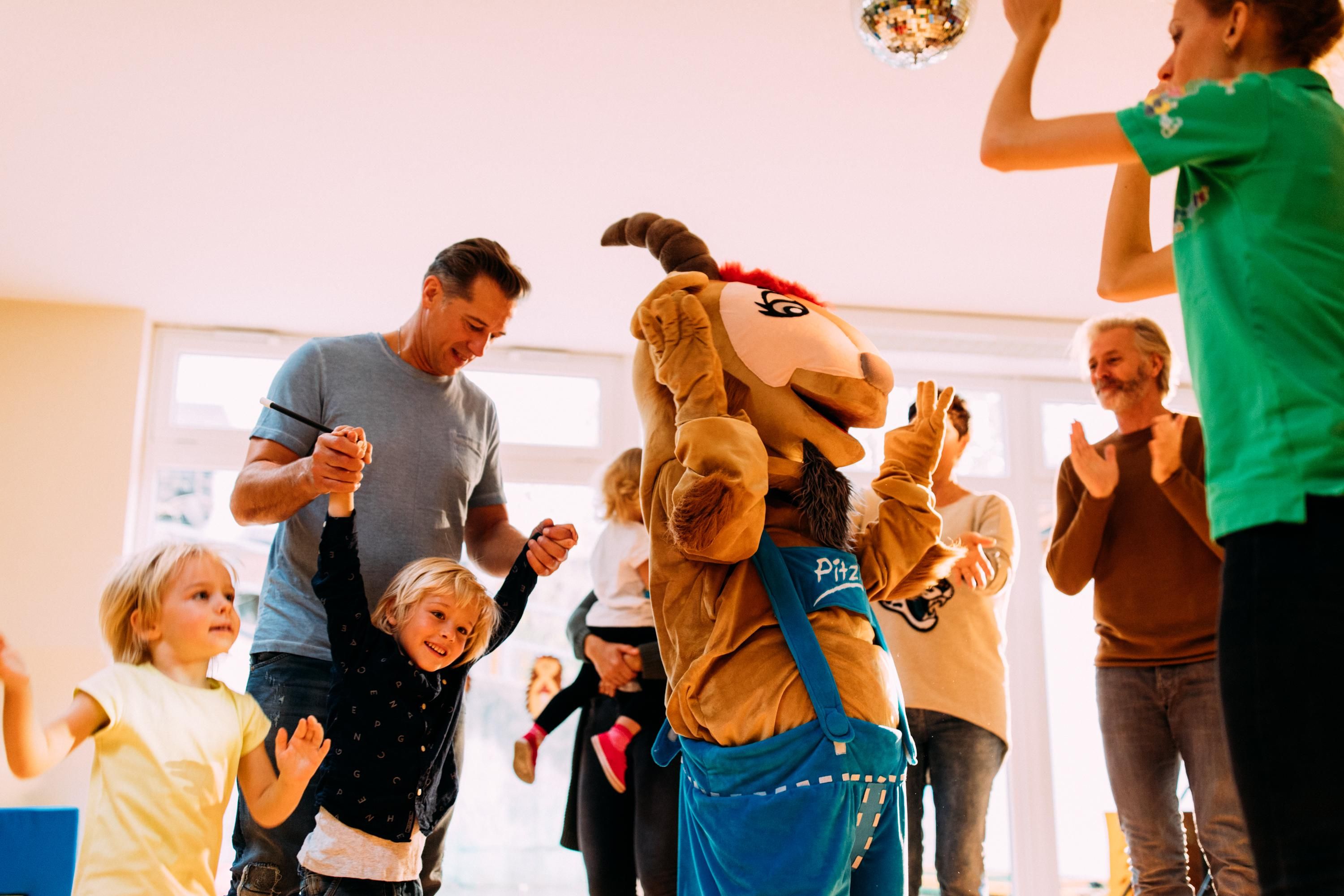 A cheerful group of children and adults are dancing in a bright room. A mascot with antlers is in the middle of the action.