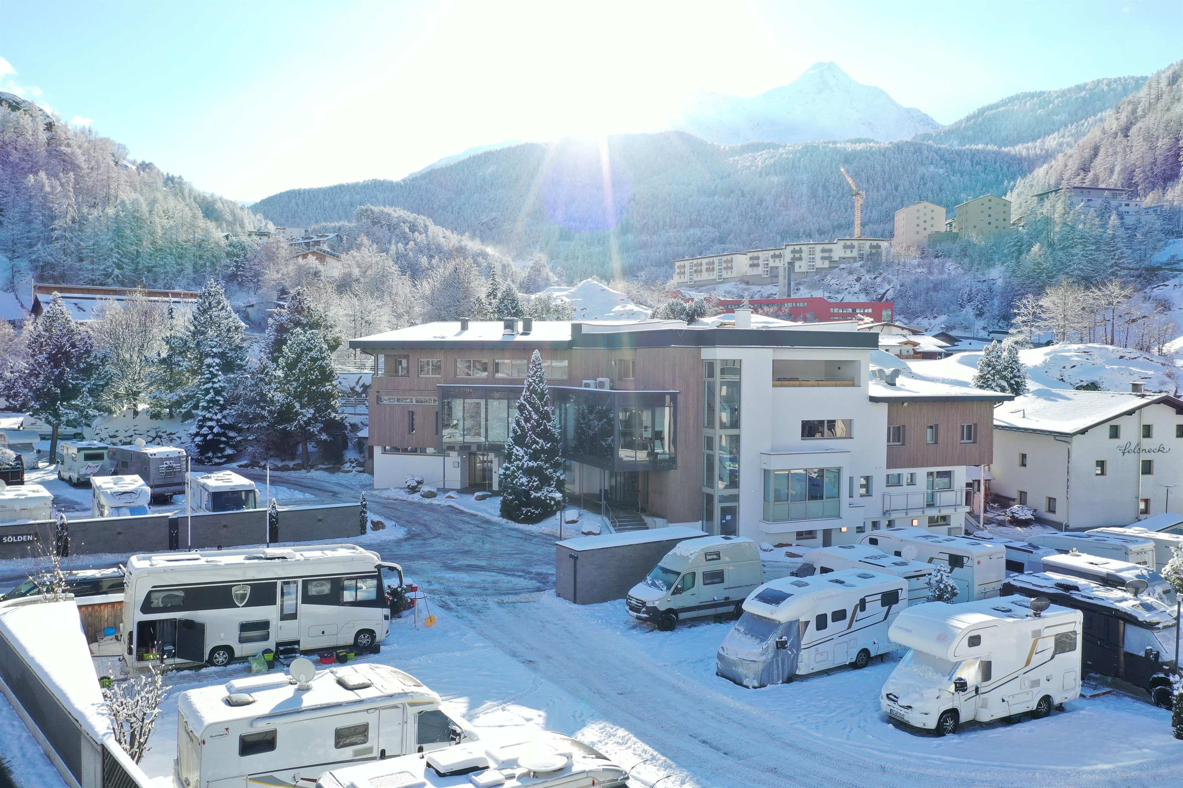 A snowy landscape with modern buildings and RVs. The sun is shining over the mountains in the background.