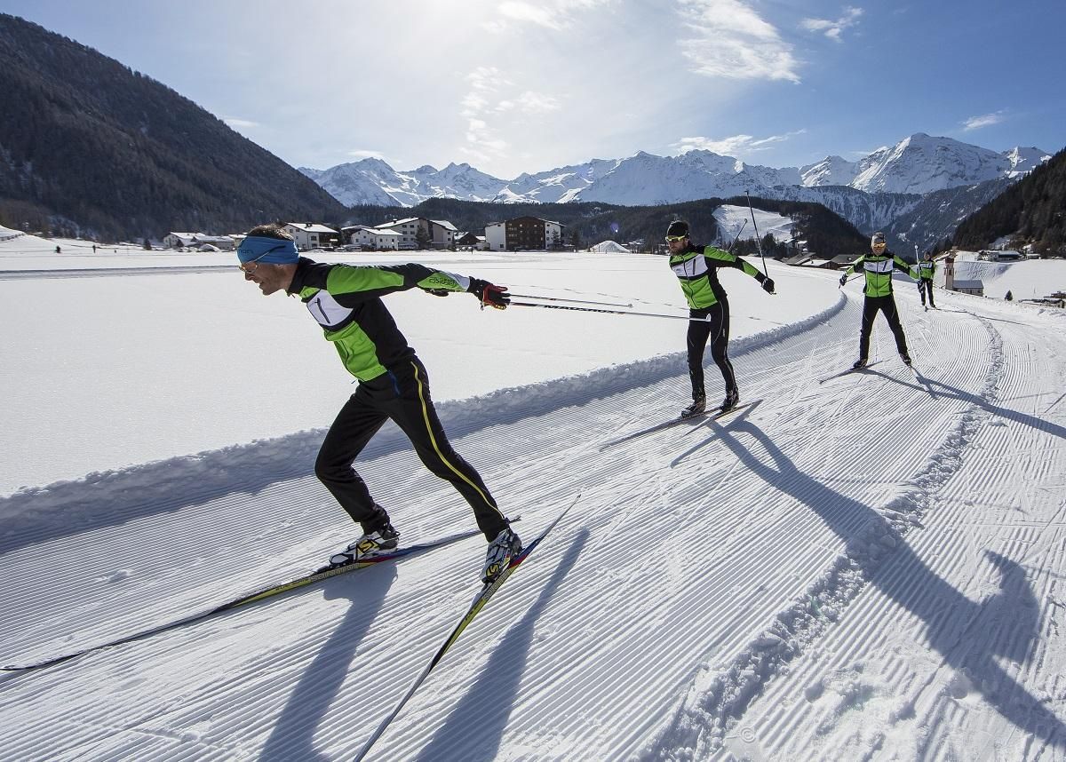 Three cross-country skiers are skating on a white, snow-covered track in the mountains. The sun is shining and the landscape is picturesque.