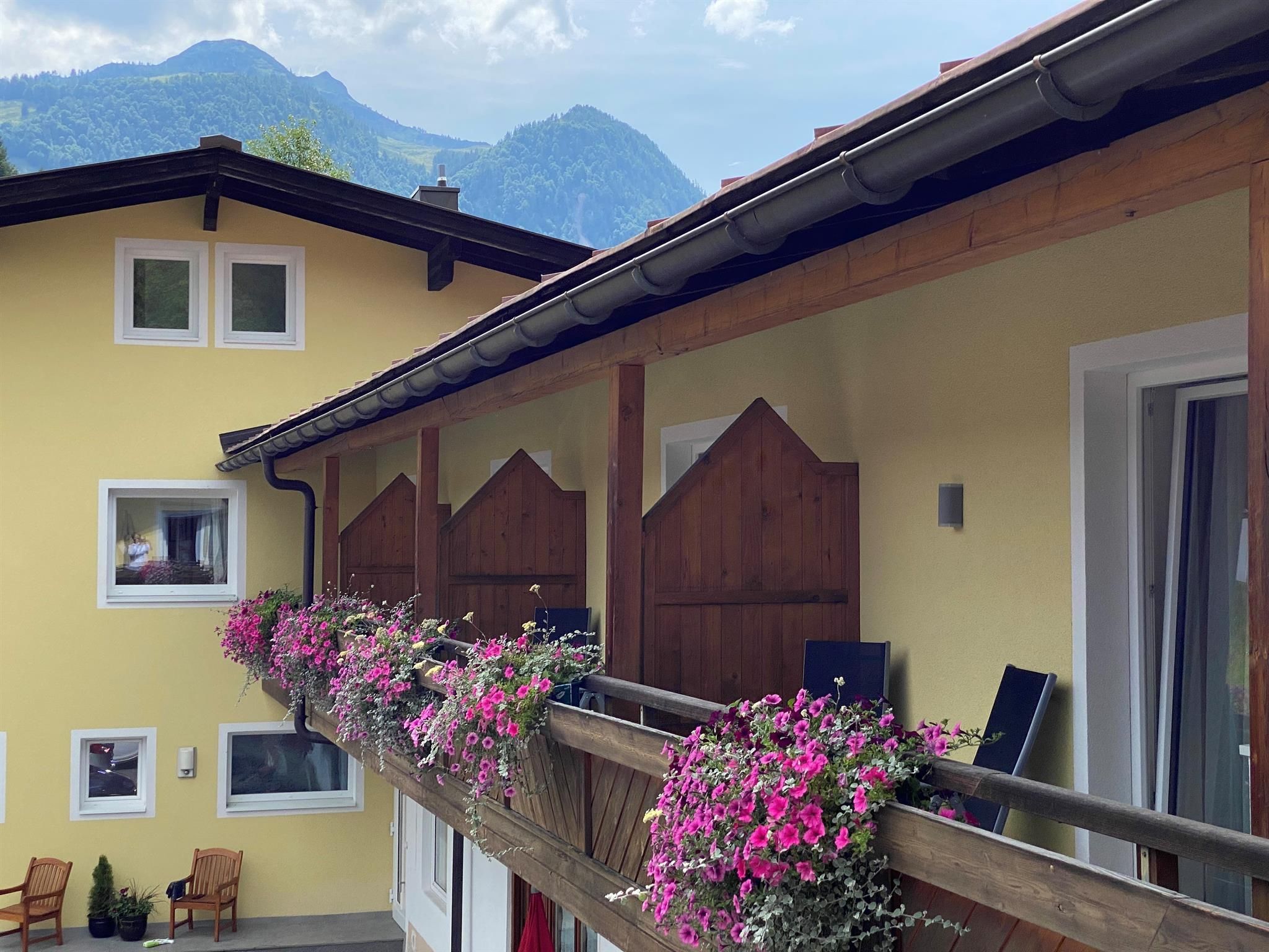 A balcony with colorful flowers and cozy chairs. In the background, there are mountains and a blue sky.