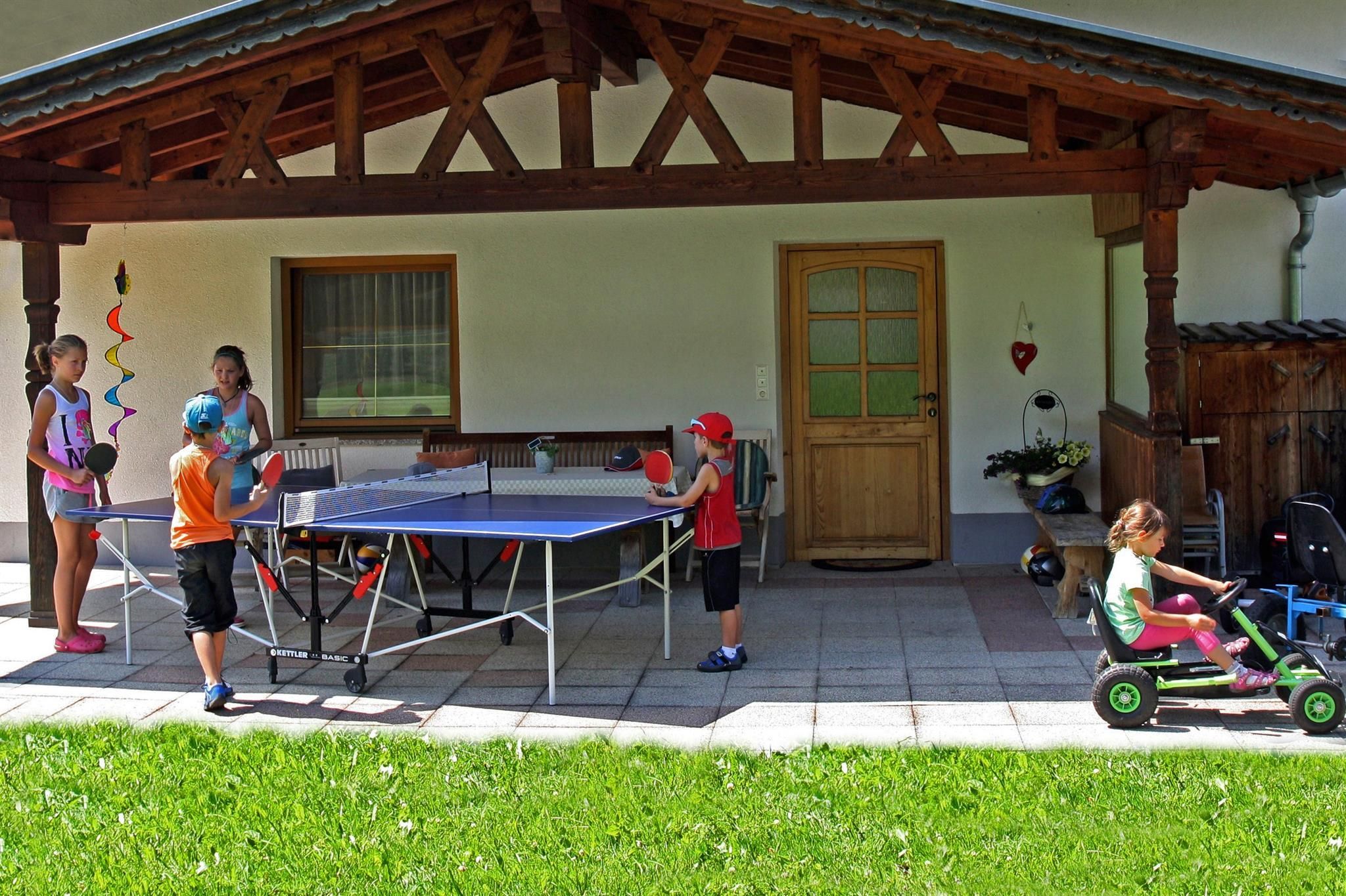 A group of children is playing table tennis on a covered terrace. Another child is riding a toy car in the grass.