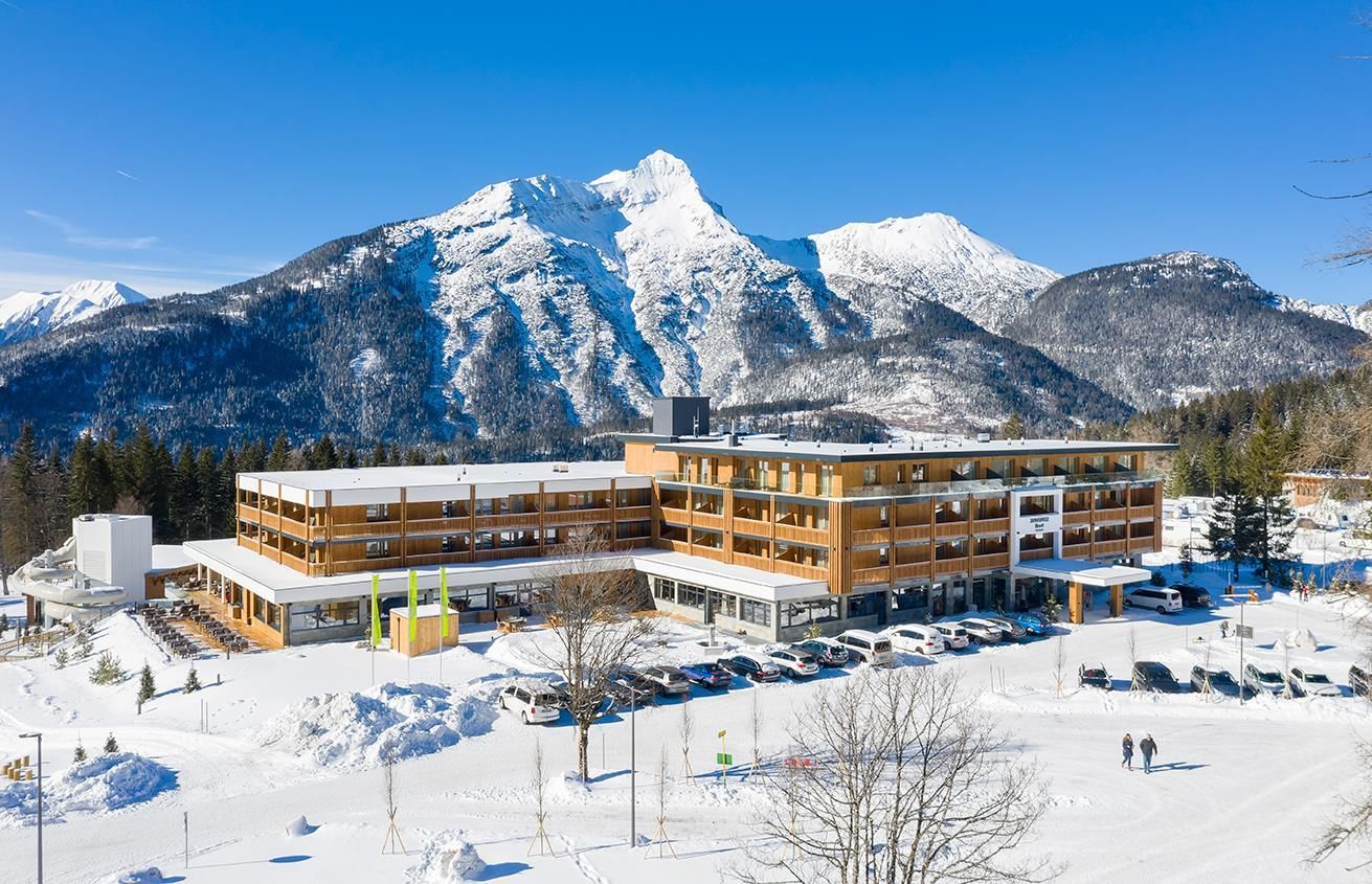 A modern hotel in a wintry landscape. In the background, snow-covered mountains rise under a blue sky.