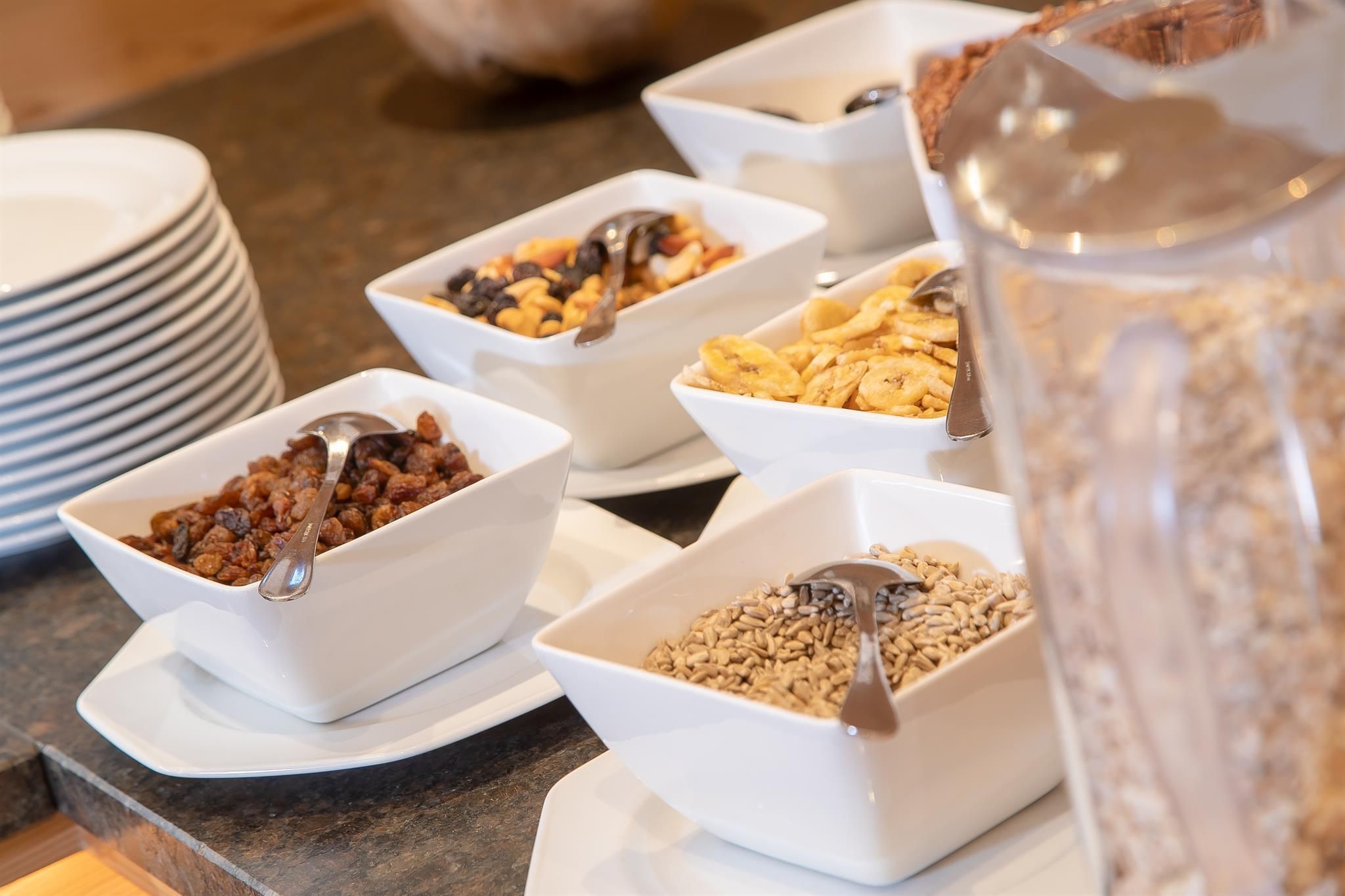 A selection of breakfast ingredients in white bowls, including muesli, dried fruits, and cornflakes. Plates are visible in the background.