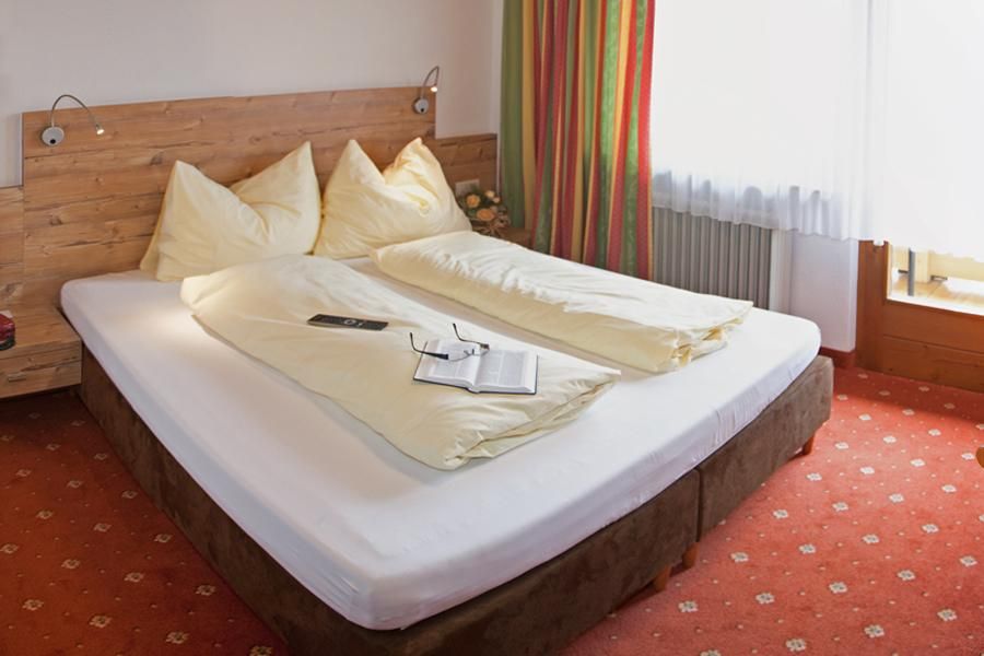 A cozy bedroom with a large bed and beige pillows. A book and a pen are on the bed, and the windows let in plenty of daylight.