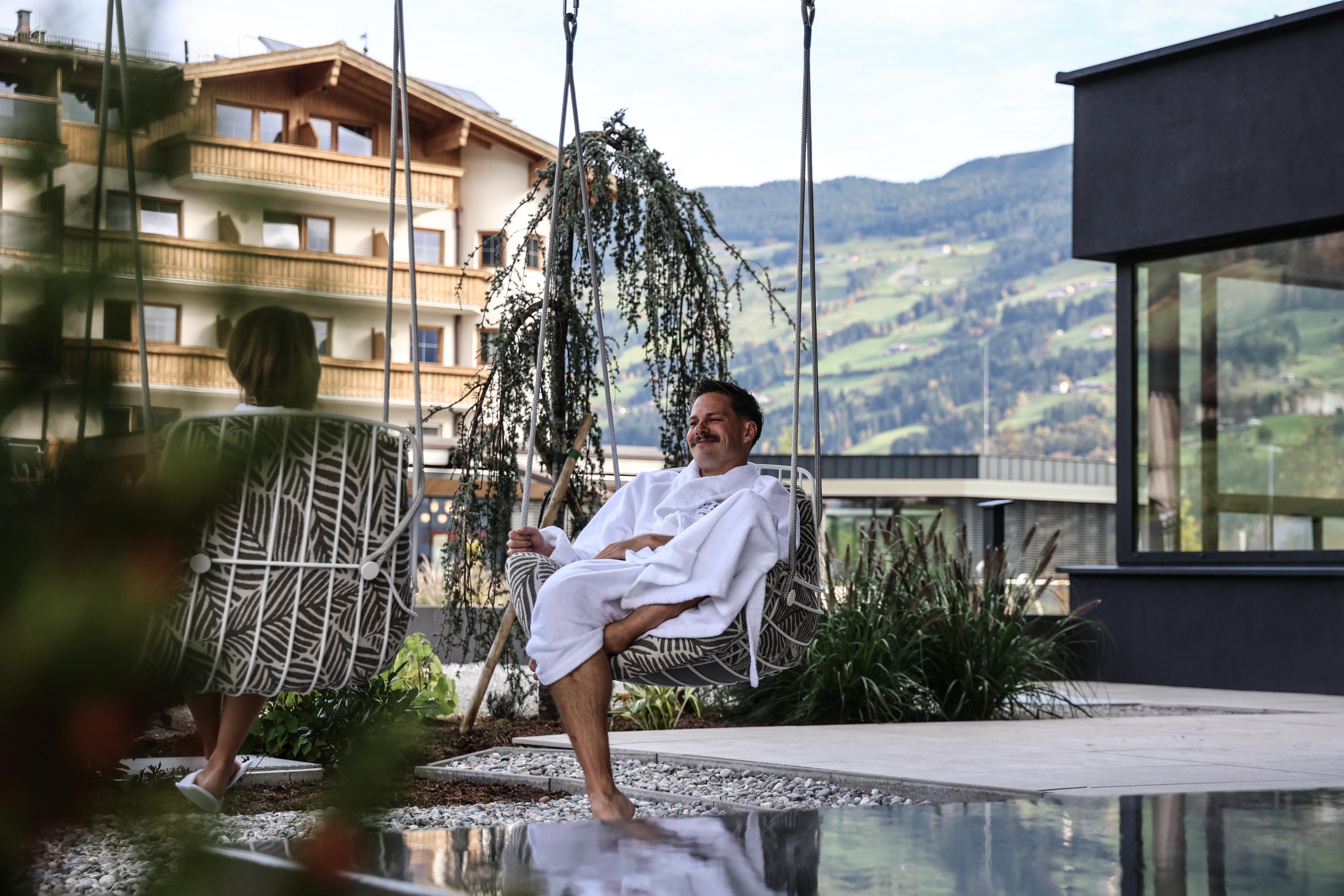 A relaxed scene outdoors, where a man is sitting in a bathrobe on a hanging swing. In the background, modern designed buildings and a green mountain landscape can be seen.