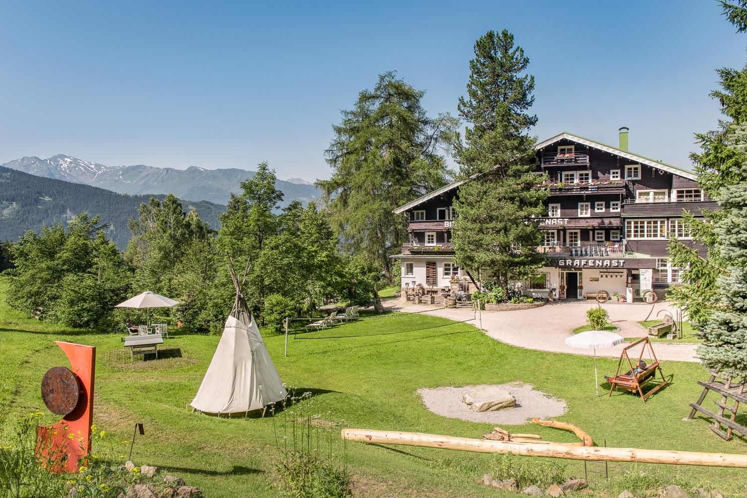 A beautiful alpine house surrounded by green meadows and trees. In the foreground, there is a tent and seating that create an inviting atmosphere.
