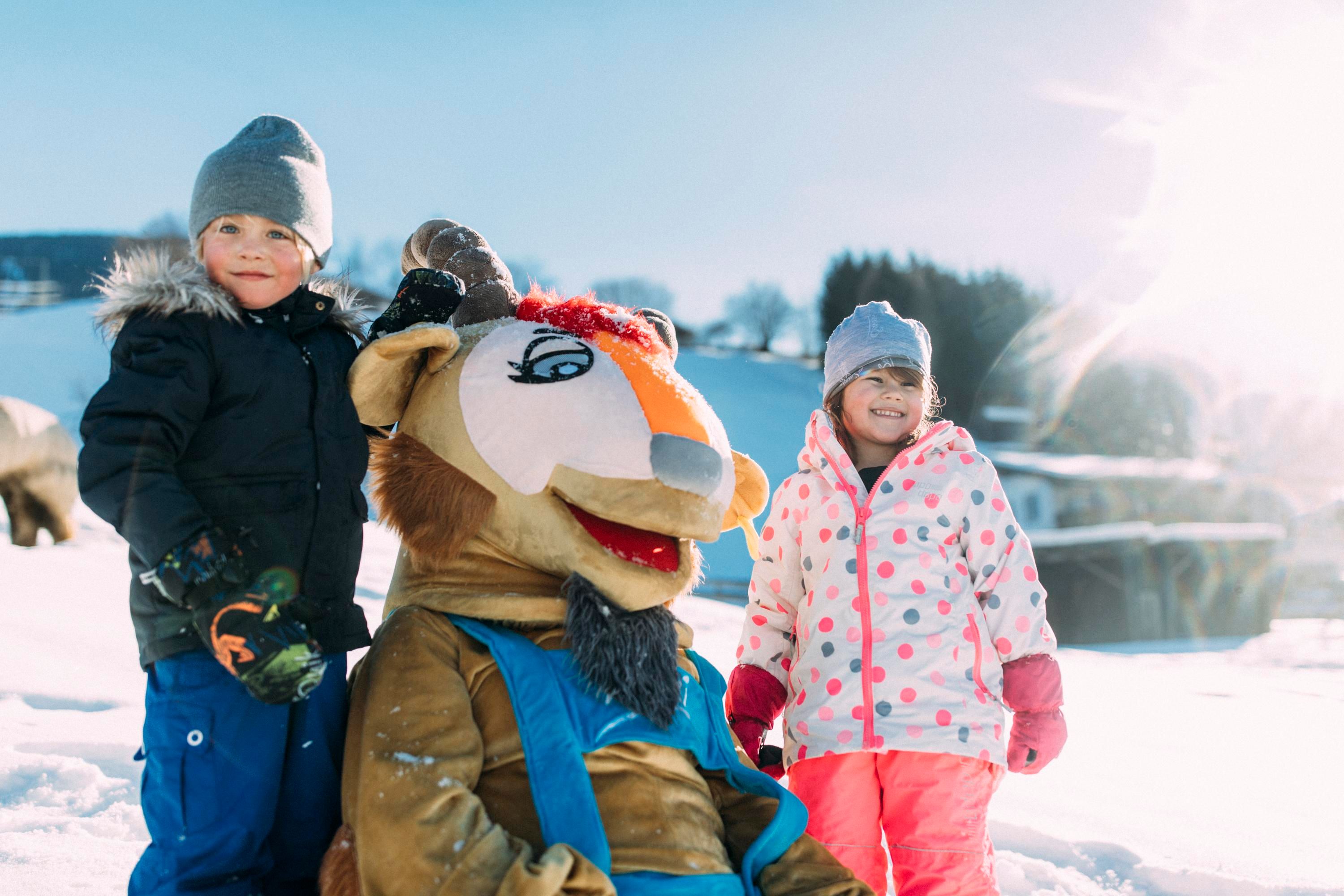 Two children are playing in the snow with a costumed animal. The sun is shining brightly and the snow is sparkling.