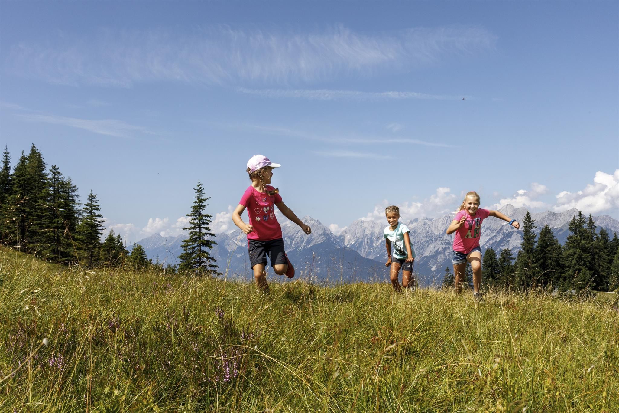 Three children are happily running across a green meadow. In the background, there are mountains and a blue sky.