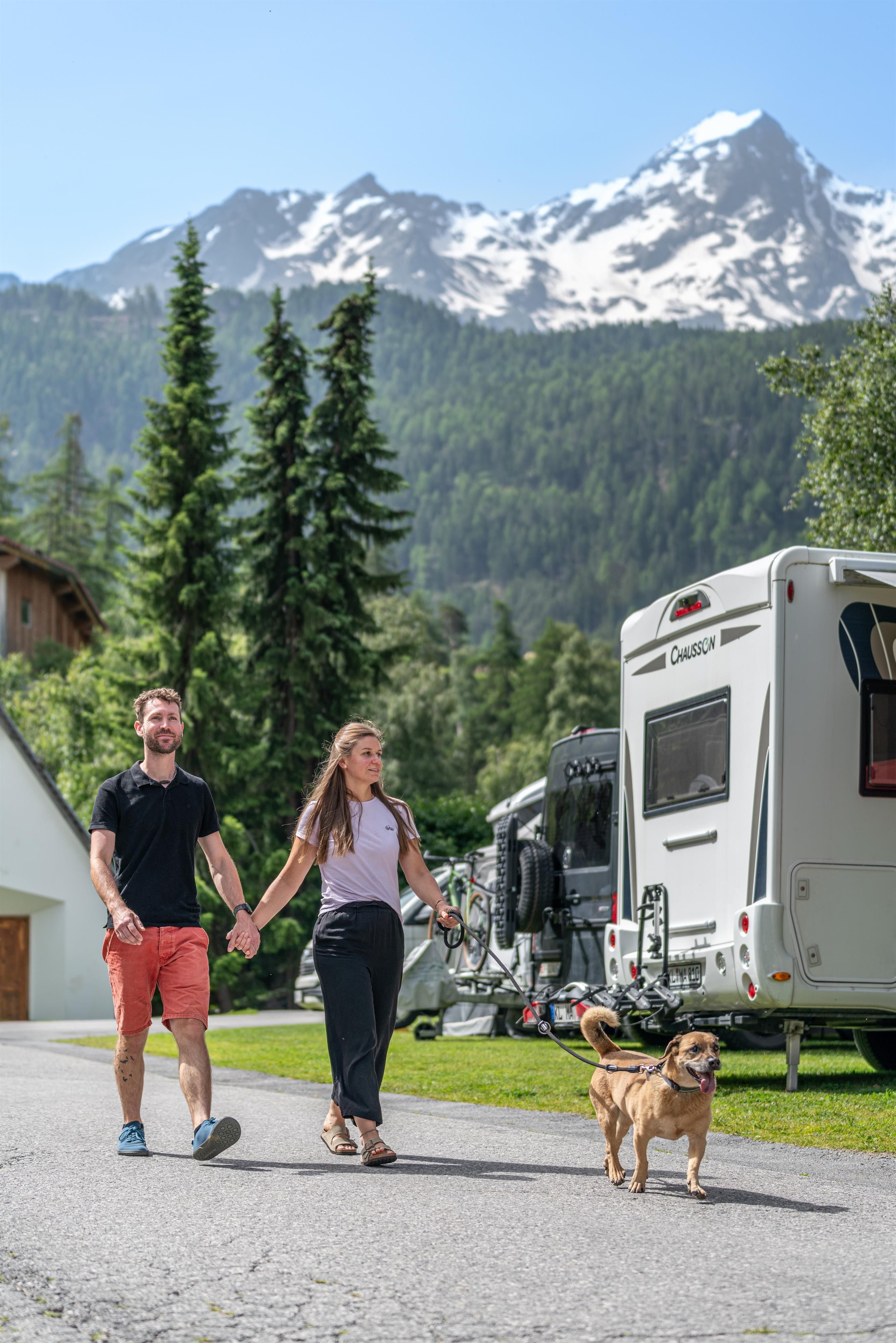 A couple is walking together with their dog on a path surrounded by nature and mountains. In the background, there are campervans and tall trees visible.