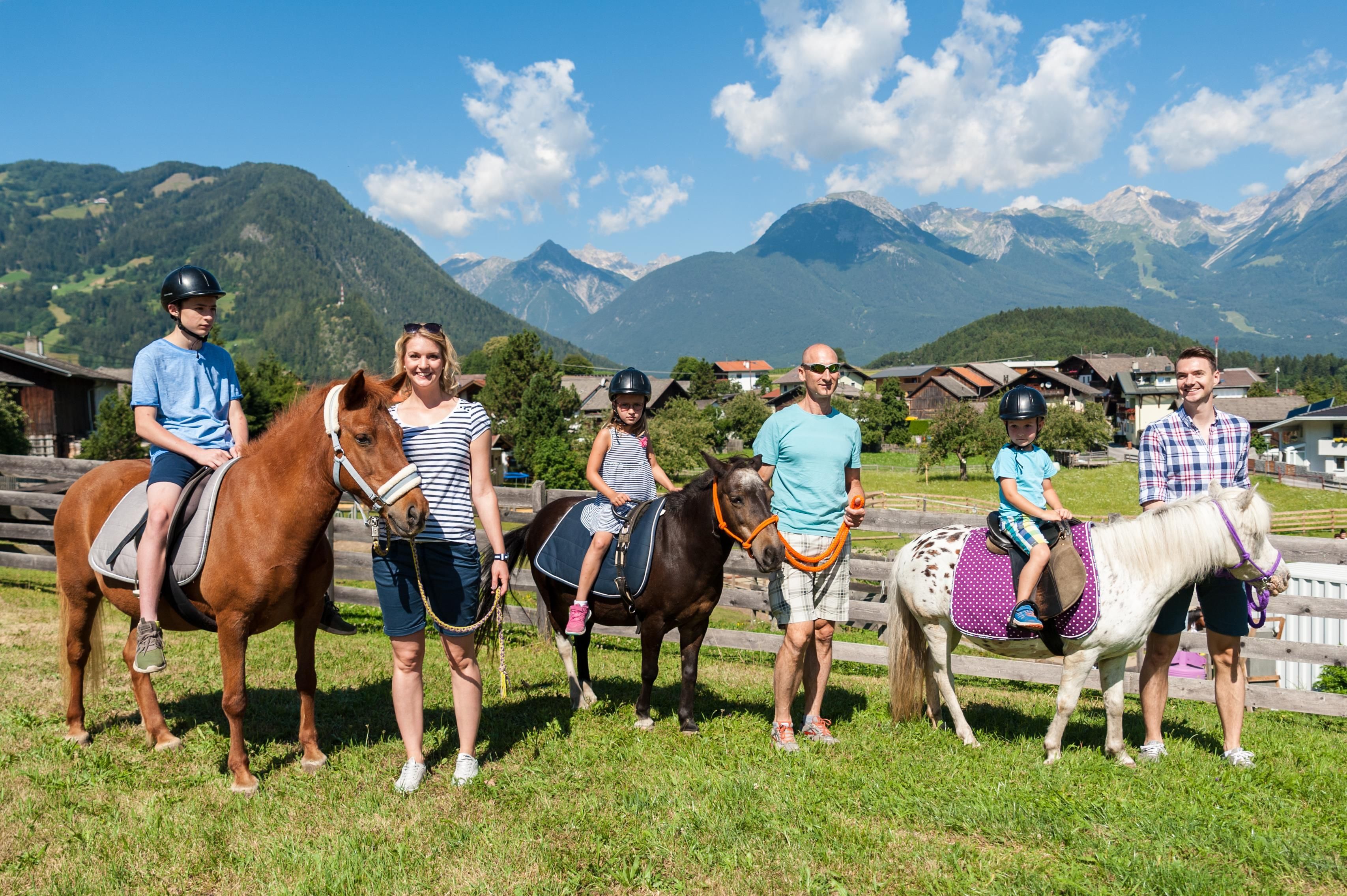 A family stands with ponies and horses in a beautiful landscape. In the background, mountains and a blue sky can be seen.