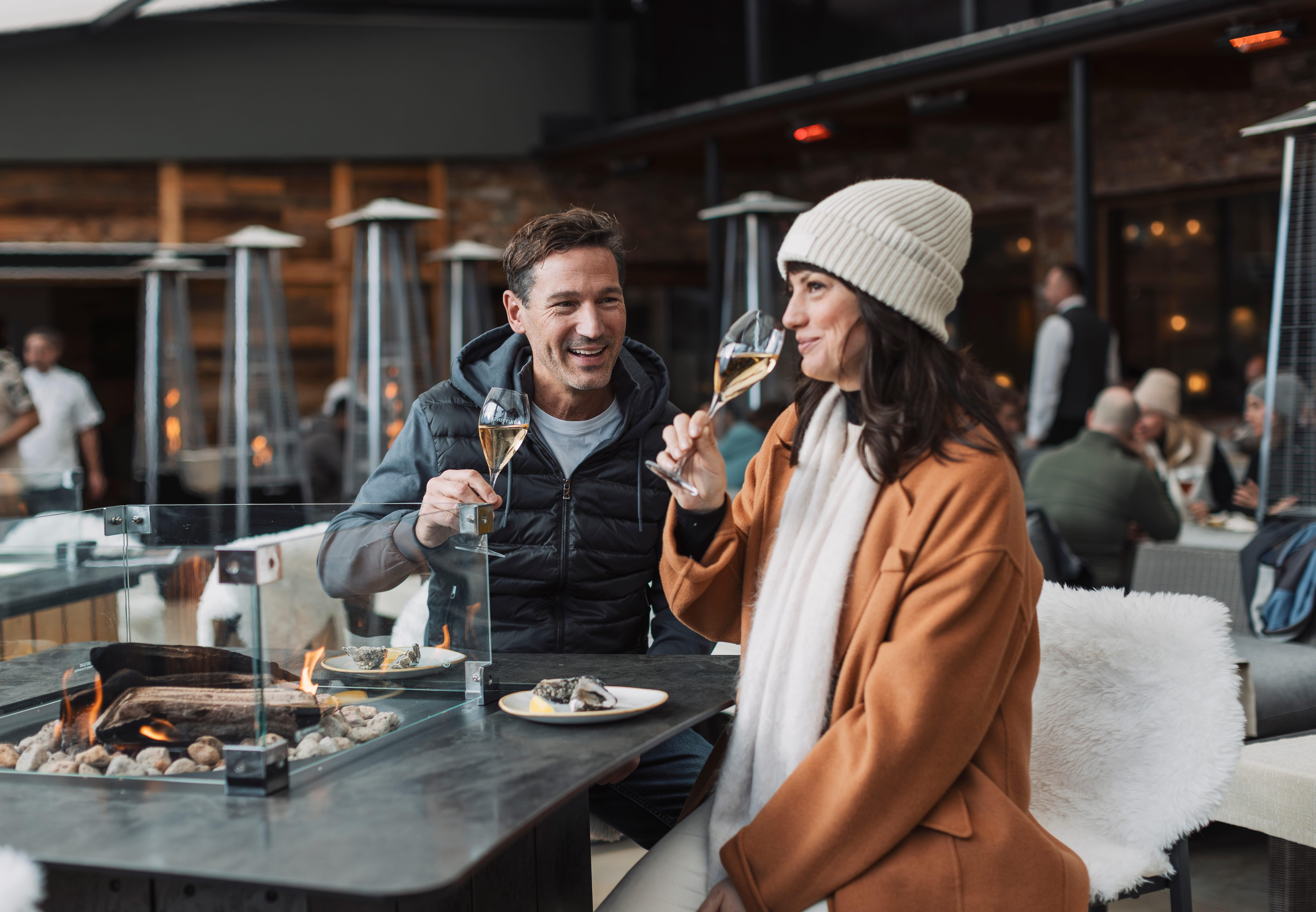 A couple enjoys champagne and small bites at a modern table. The surroundings feel cozy and inviting with a festive atmosphere.