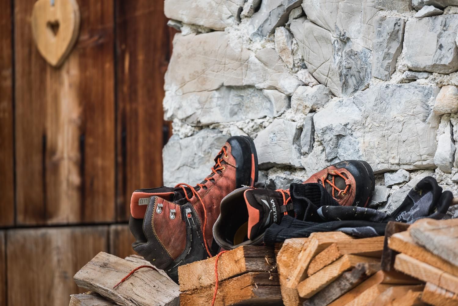 Boots and clothing are lying on a wooden stack next to a stone wall. In the background, a wooden door can be seen.