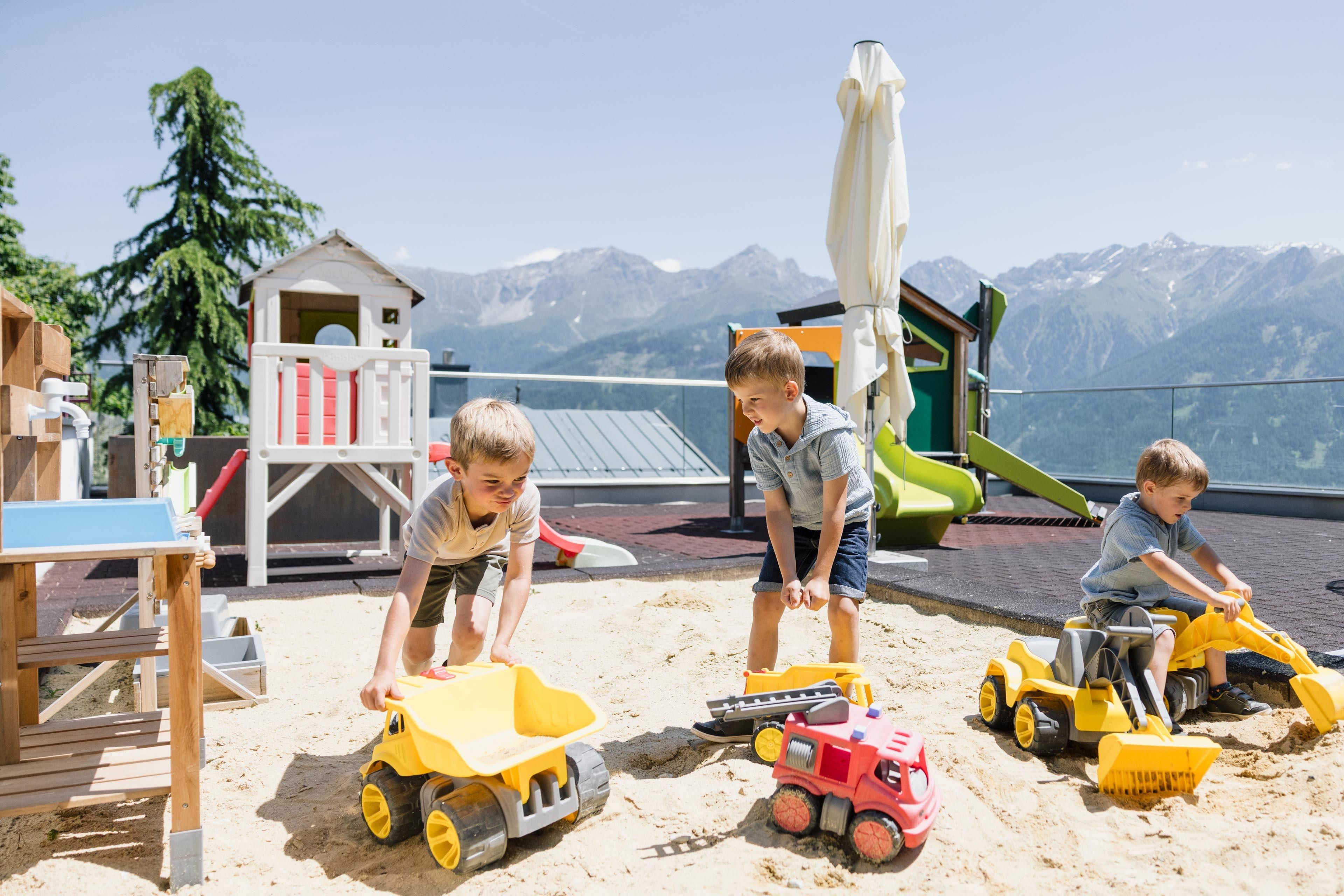 Children are playing in the sand with construction vehicles on a terrace. In the background, a colorful playhouse and other play equipment can be seen. Additionally, there is a breathtaking view of the mountains.
