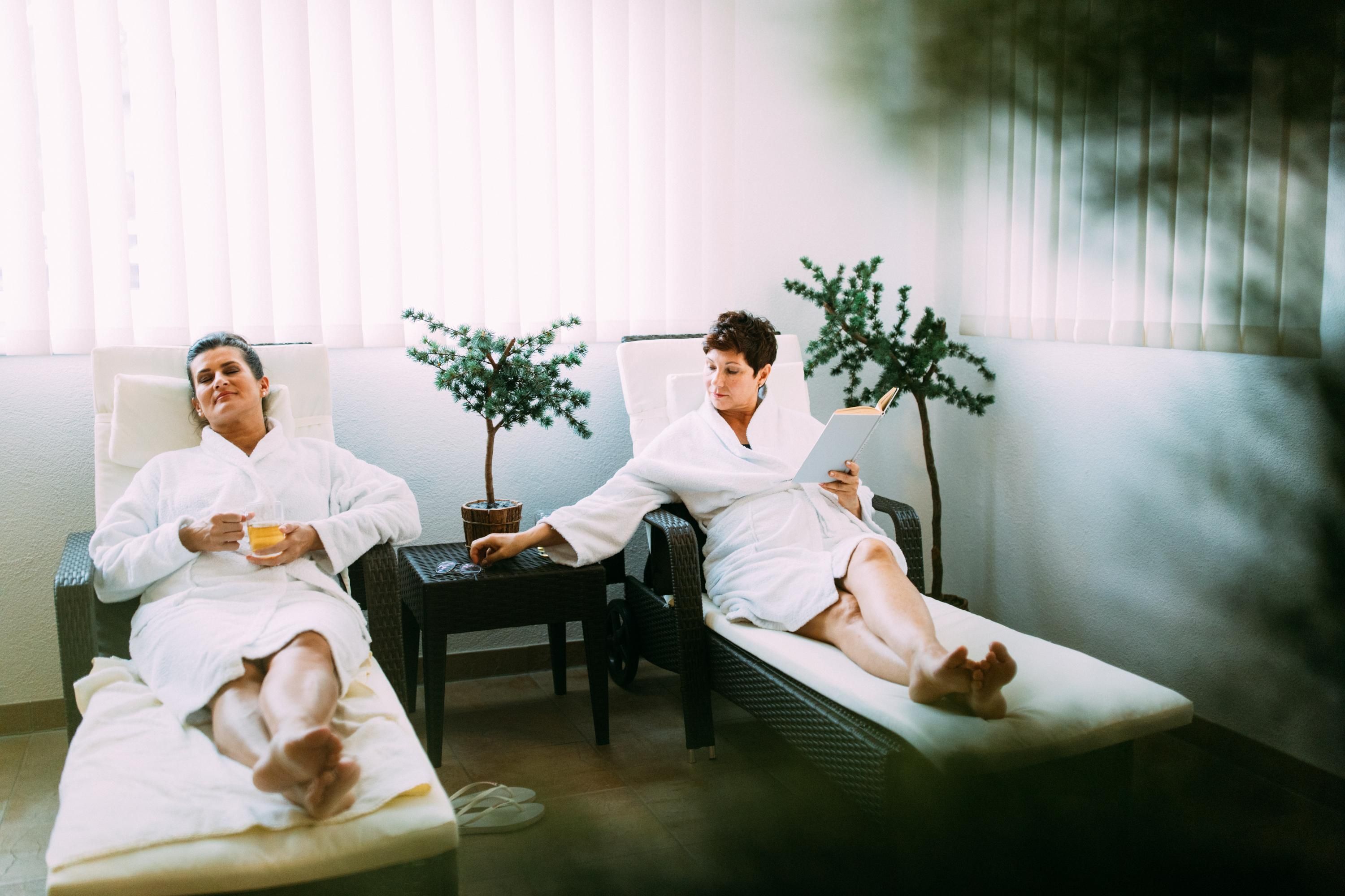 Two people in bathrobes relax on loungers in a spa. The room is brightly lit, with plants in the background.