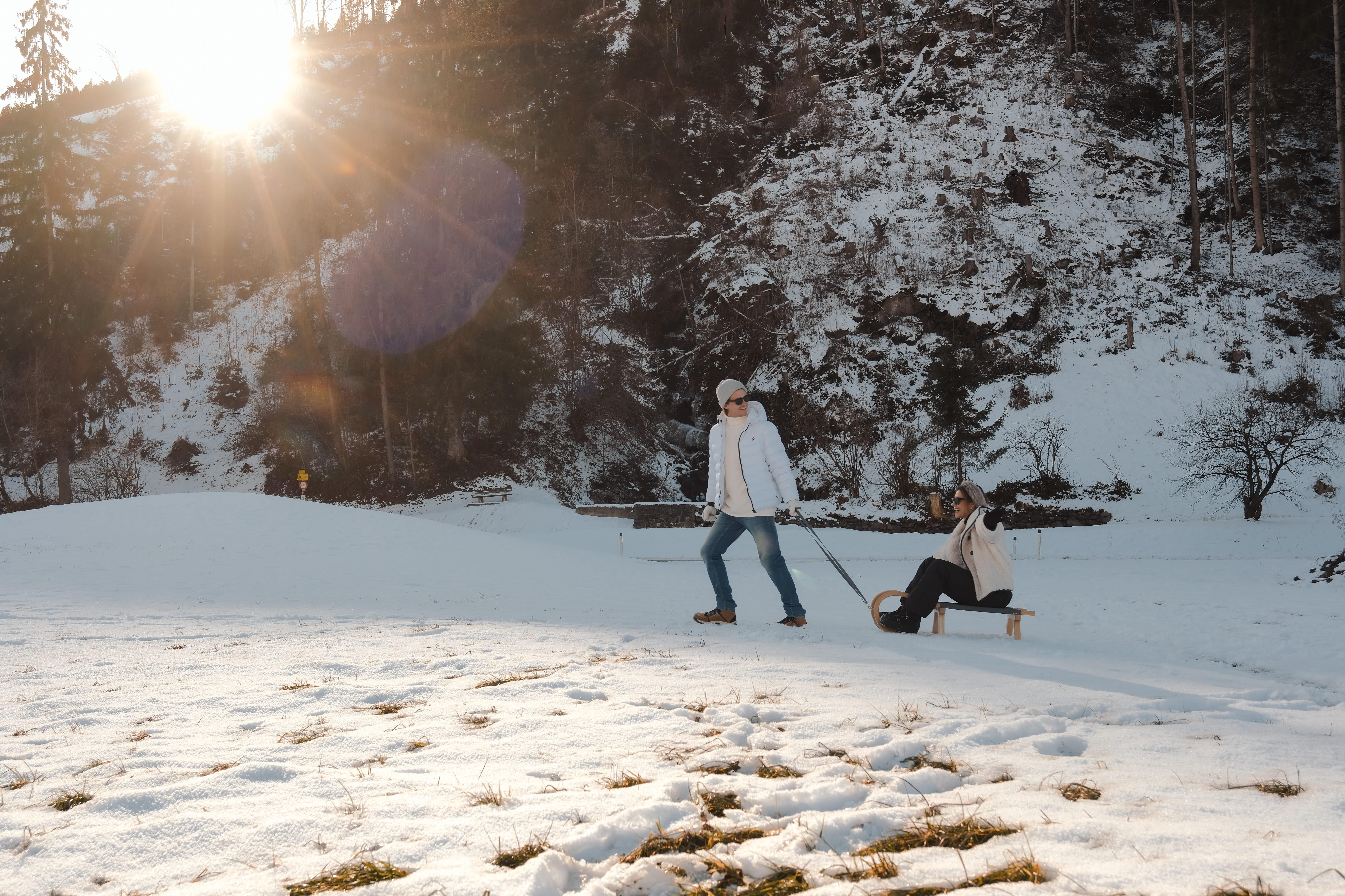 A wintry landscape with snow and trees in the background. One person is pulling a sled on which another person is sitting.
