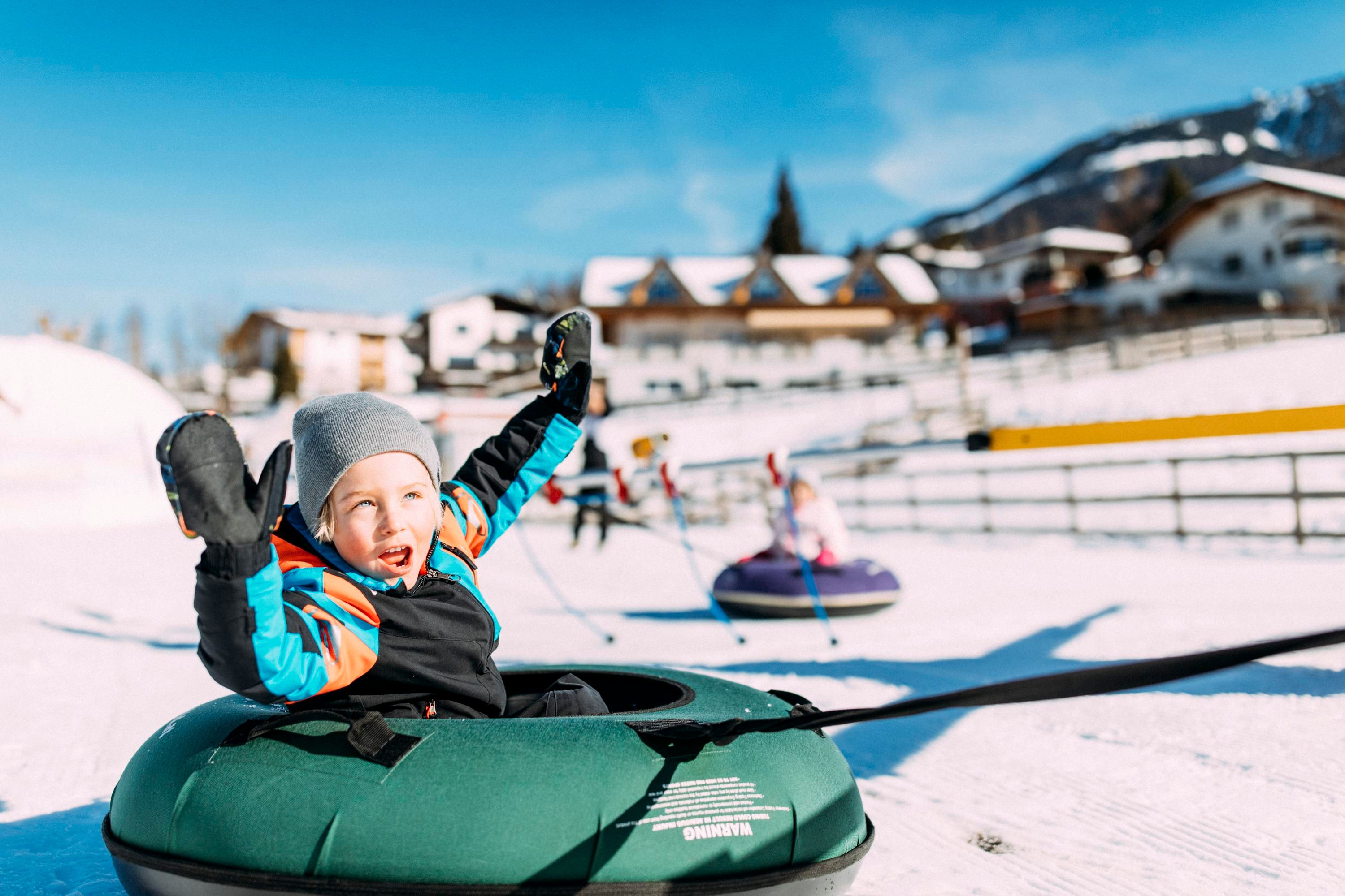 A happy child is sliding down the hill in a snow tubing tire. In the background, mountains and houses can be seen.