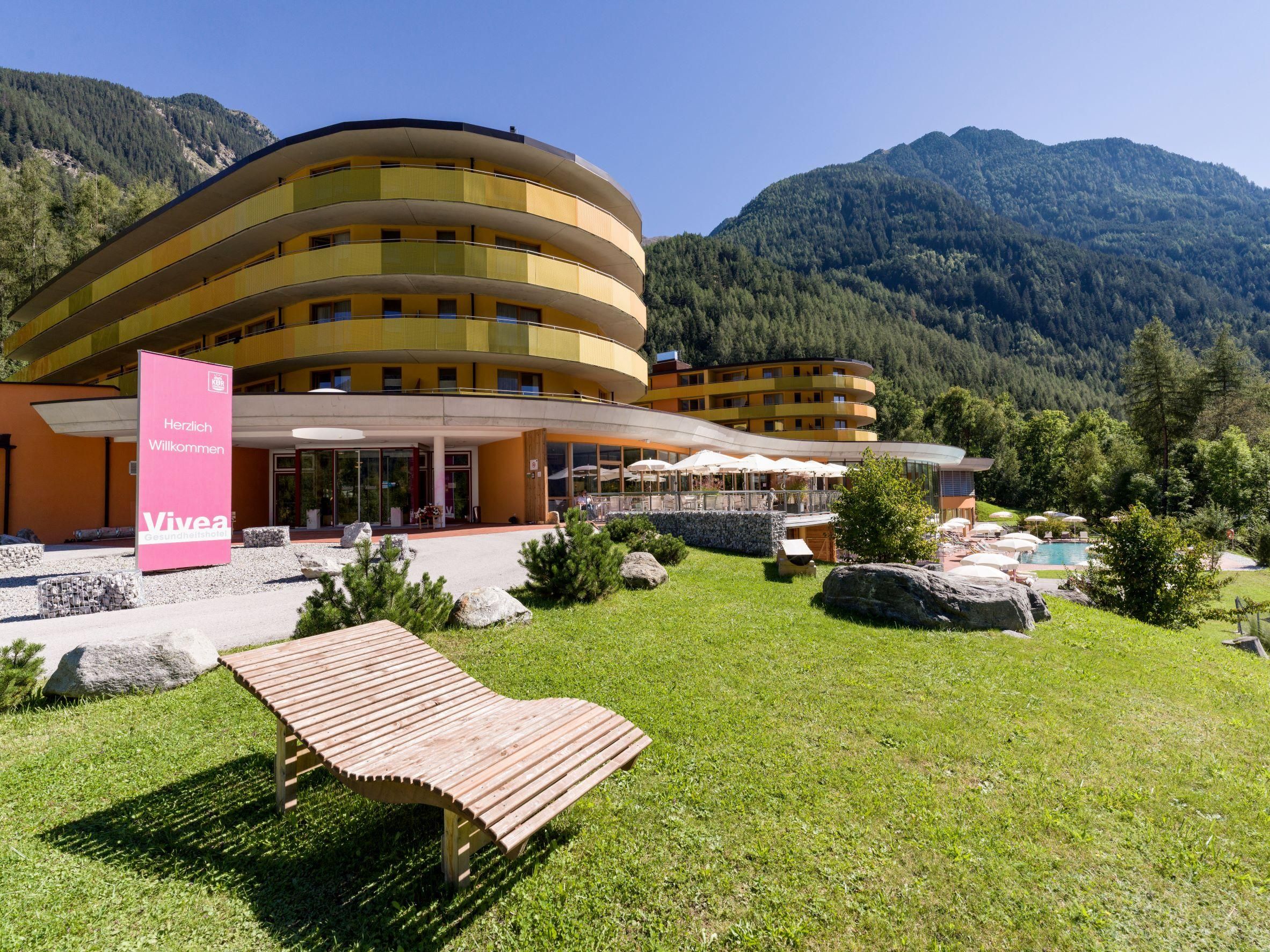 A modern hotel amidst green nature and mountains. The building has a yellow facade and offers an inviting terrace with loungers.