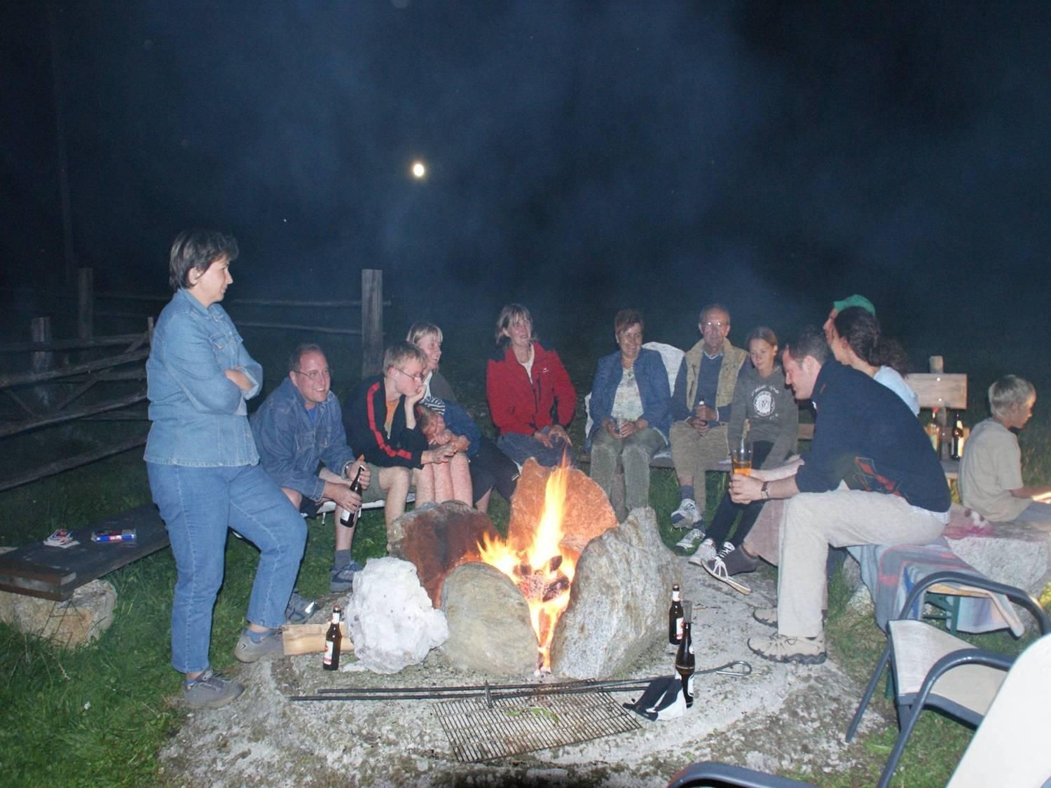 A group of people is sitting around a campfire in a nighttime setting. The sky is dark, and the moon is visible.