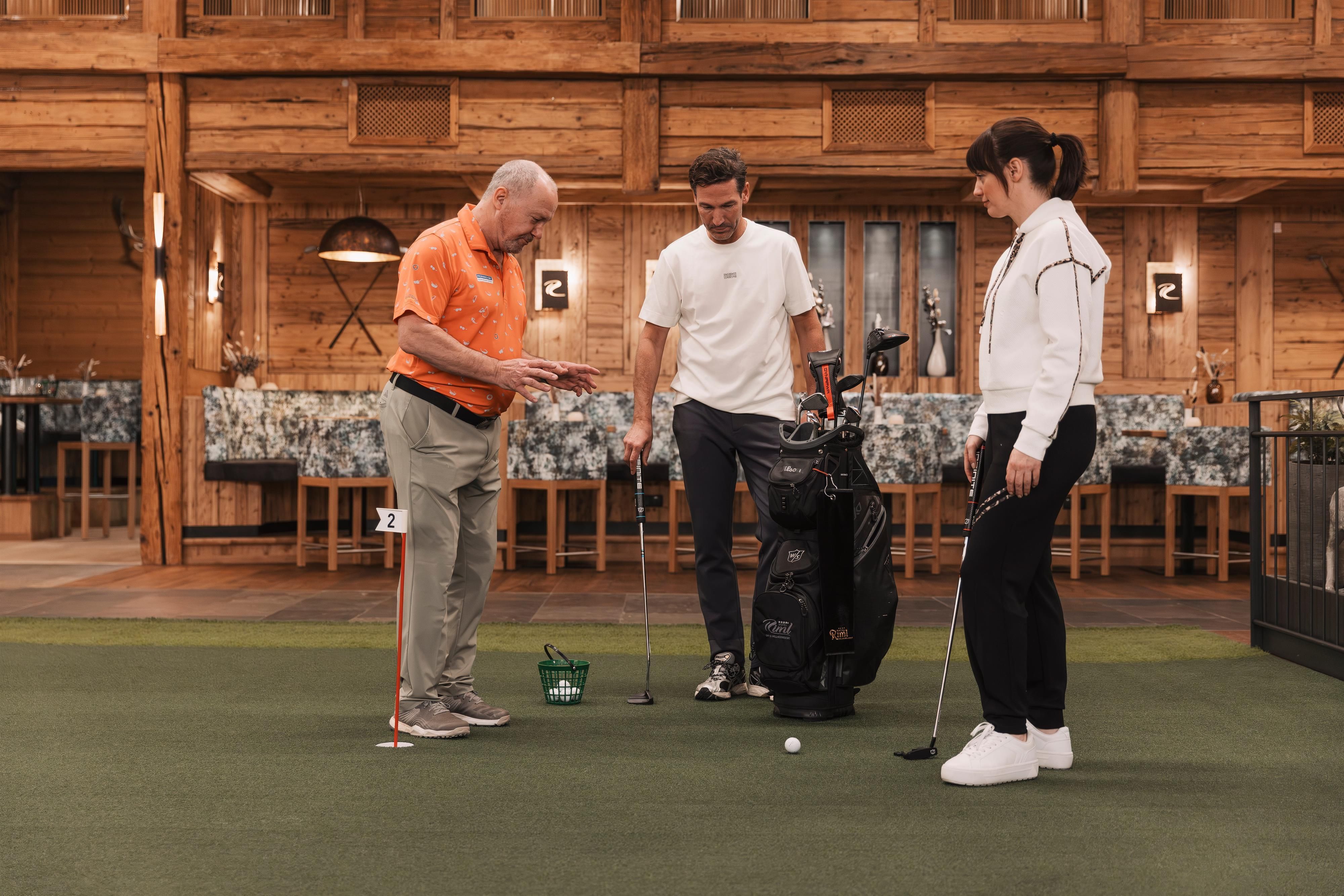 A golf instructor is showing two people how to putt. They are in a wood-paneled indoor golf environment.