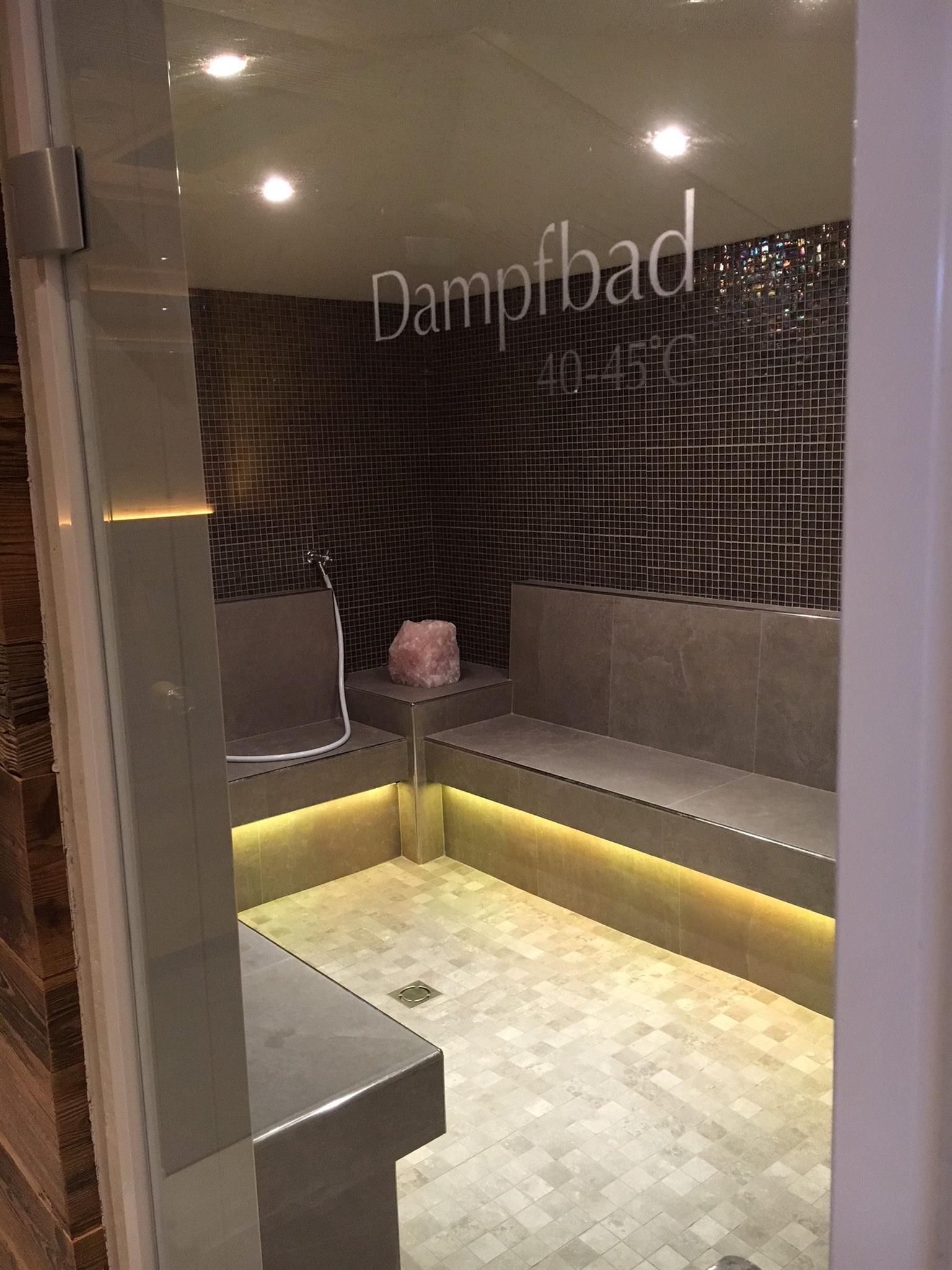 A modern steam bath with gray benches and indirect lighting. The walls are lined with tiles and there is a faucet for use.
