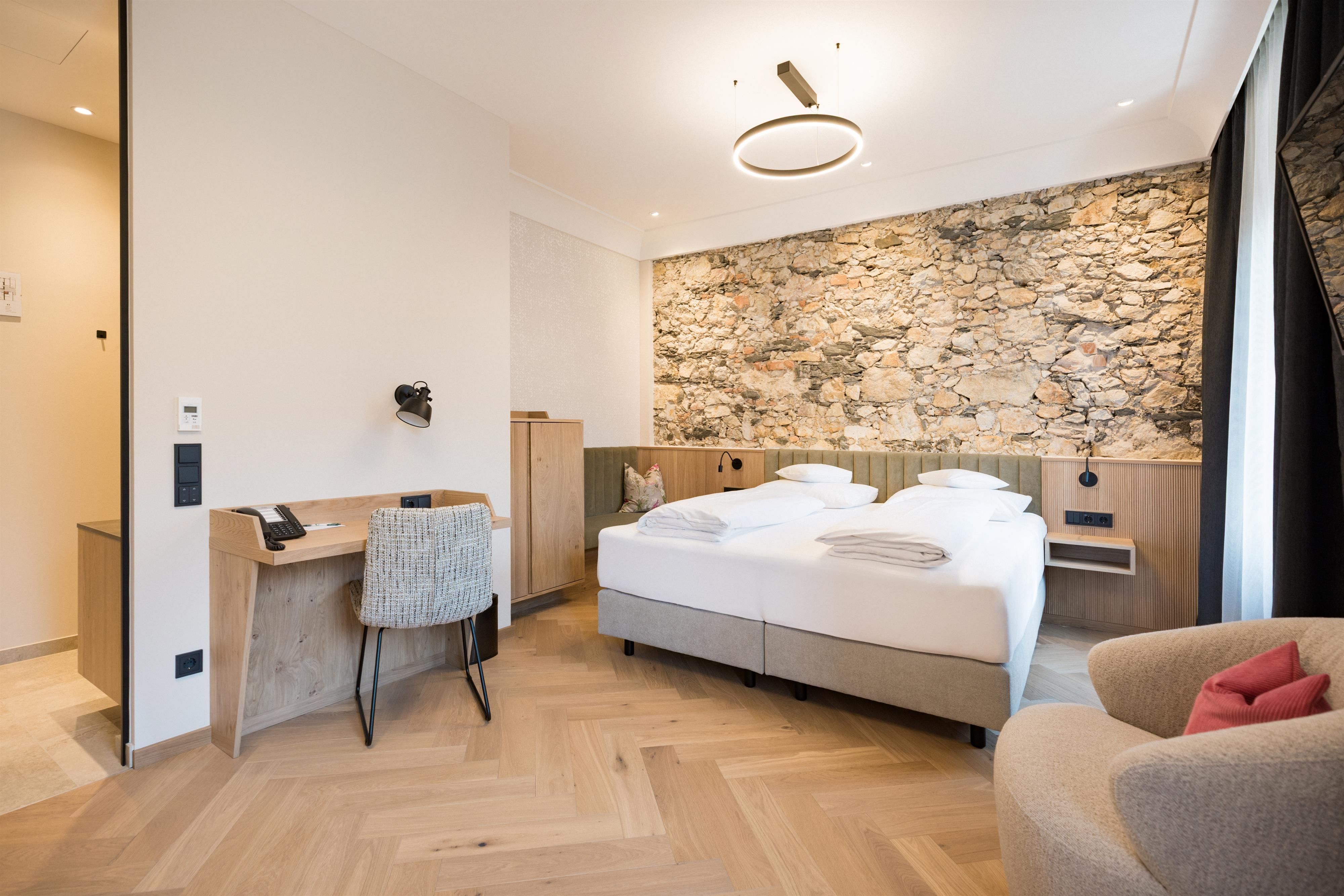 A modern hotel room with a large bed and a stone wall. There is a desk and a cozy armchair in a bright, friendly space.