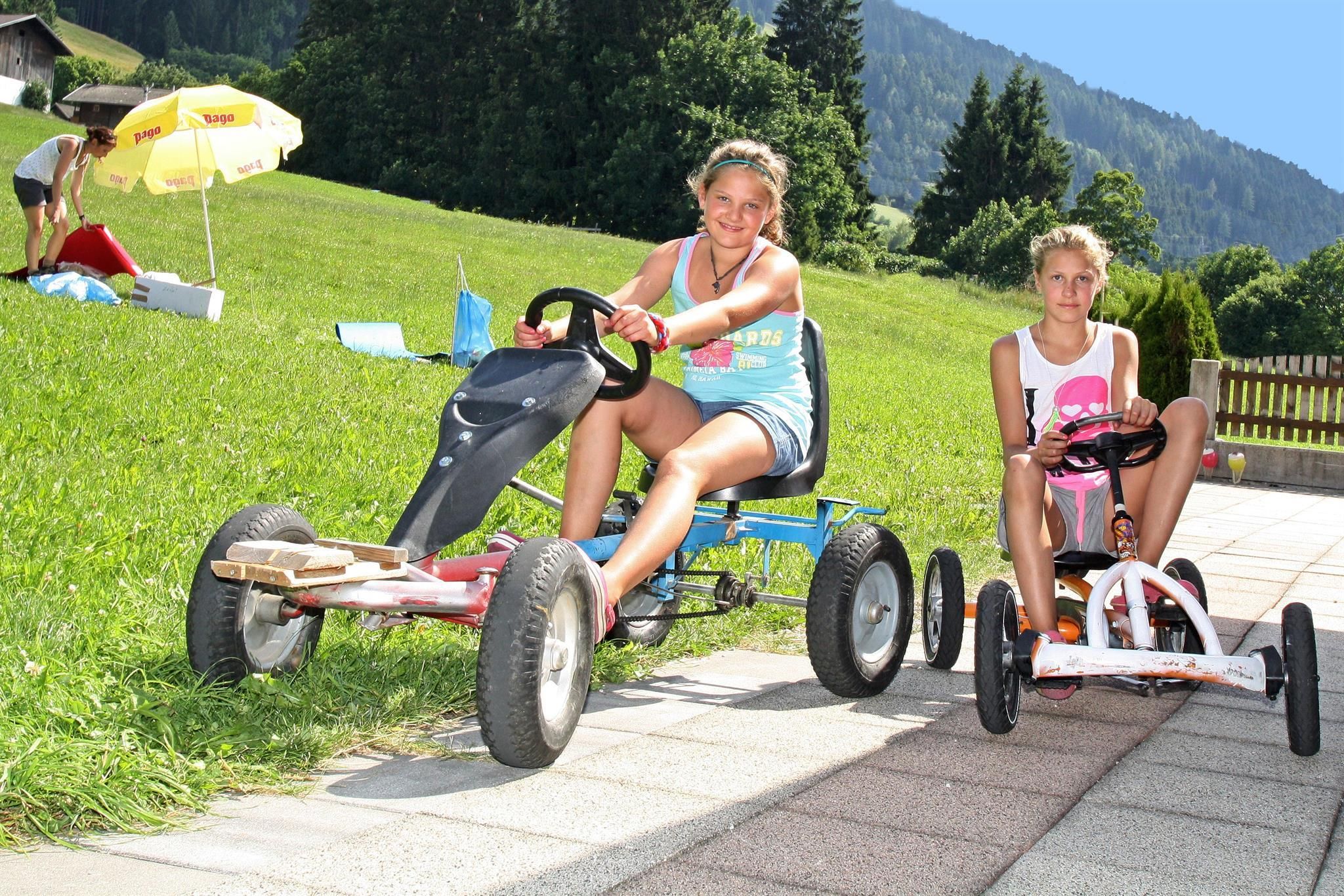 Two girls are riding go-karts on a paved path. In the background, there are fields and trees.
