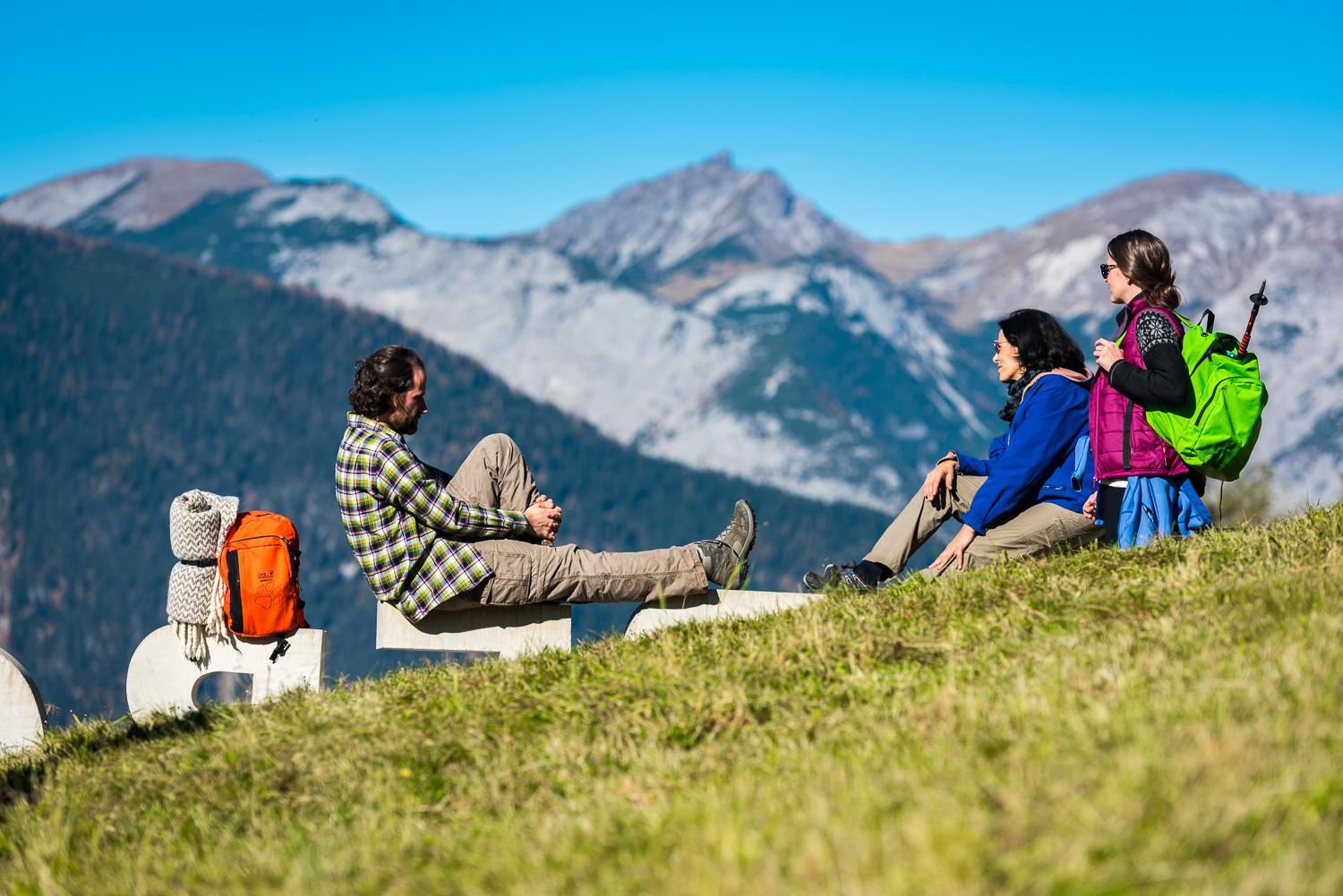 Three people are relaxing on a meadow in the mountains. In the background, green hills and snow-capped peaks can be seen.