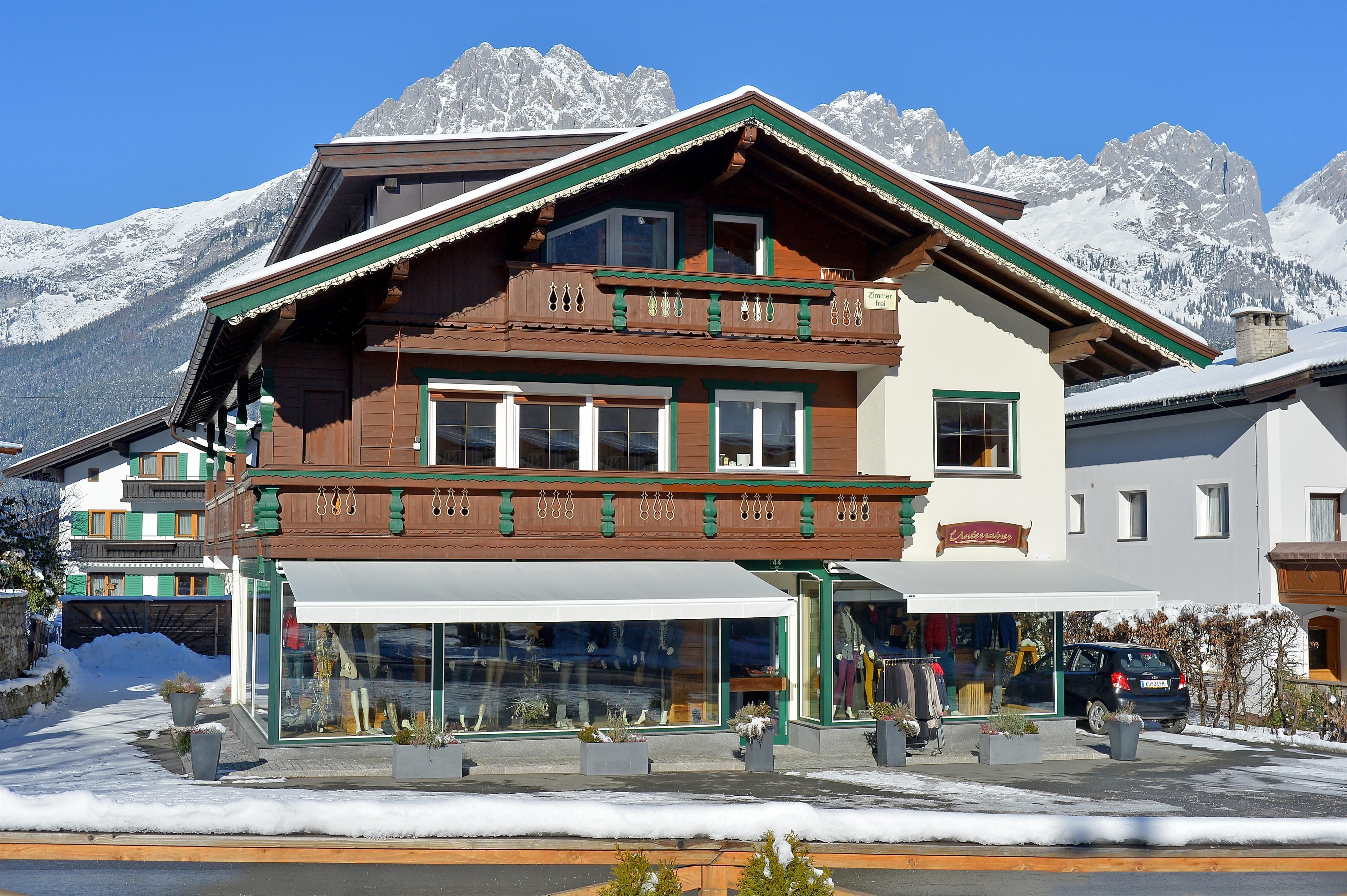A traditional alpine house with wooden cladding and a balcony. In the foreground, snow-covered areas are visible along with the impressive mountains in the background.