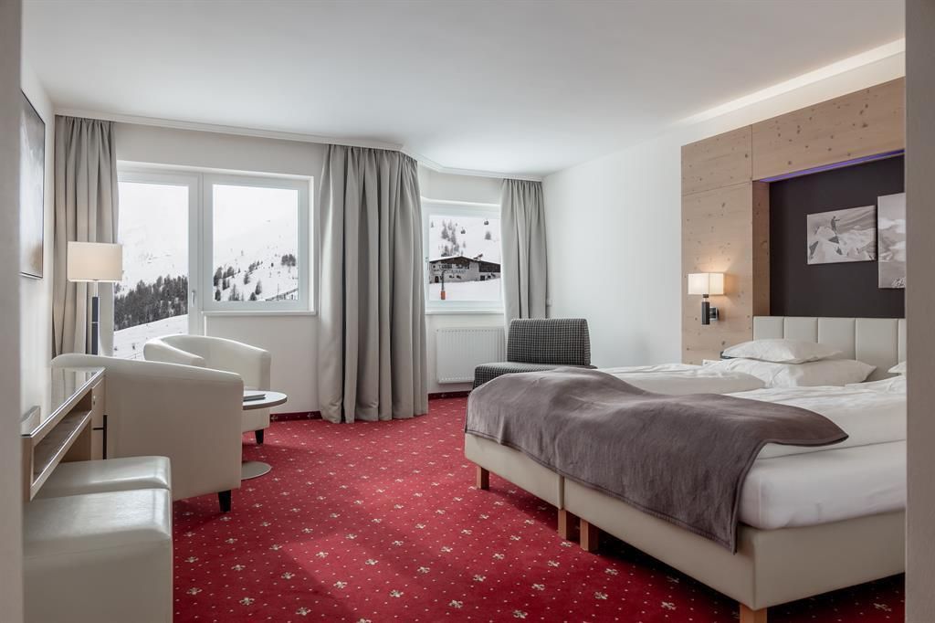 A modern hotel room with a double bed and a cozy seating area. Large windows offer a view of the snow-covered mountains.