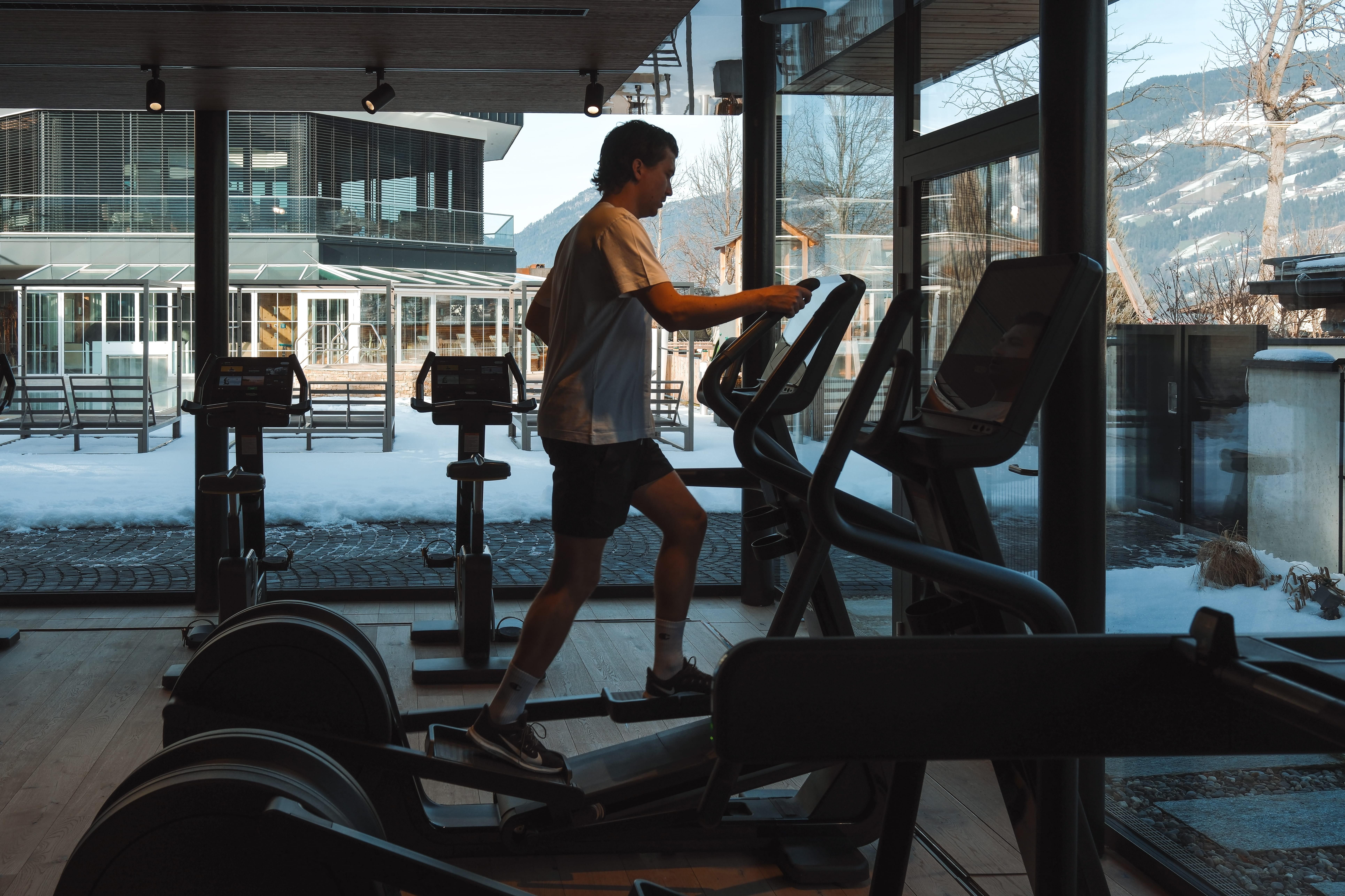 A young man is training on an elliptical trainer in a modern gym. Through the windows, snow-covered landscapes and mountains are visible.