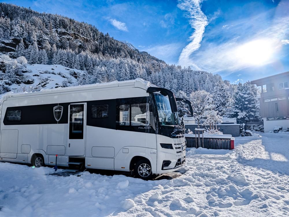A modern motorhome stands in a snow-covered landscape. In the background, there are snowy trees and a clear blue sky.
